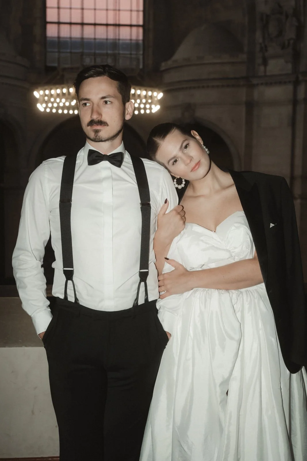 Ein Mann in Smoking und eine Frau in einem weißen Kleid posieren in einem edlen, dunklen Raum.