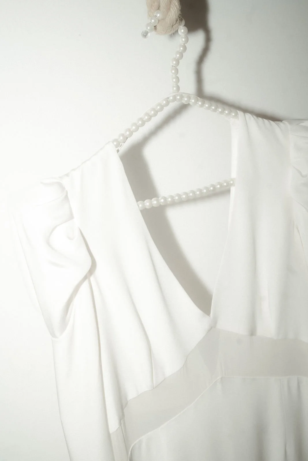 Weiße Bluse mit Rüschen an den Ärmeln, auf einem Kleiderbügel mit Perlenschnur. Hintergrund ist eine weiße Wand.