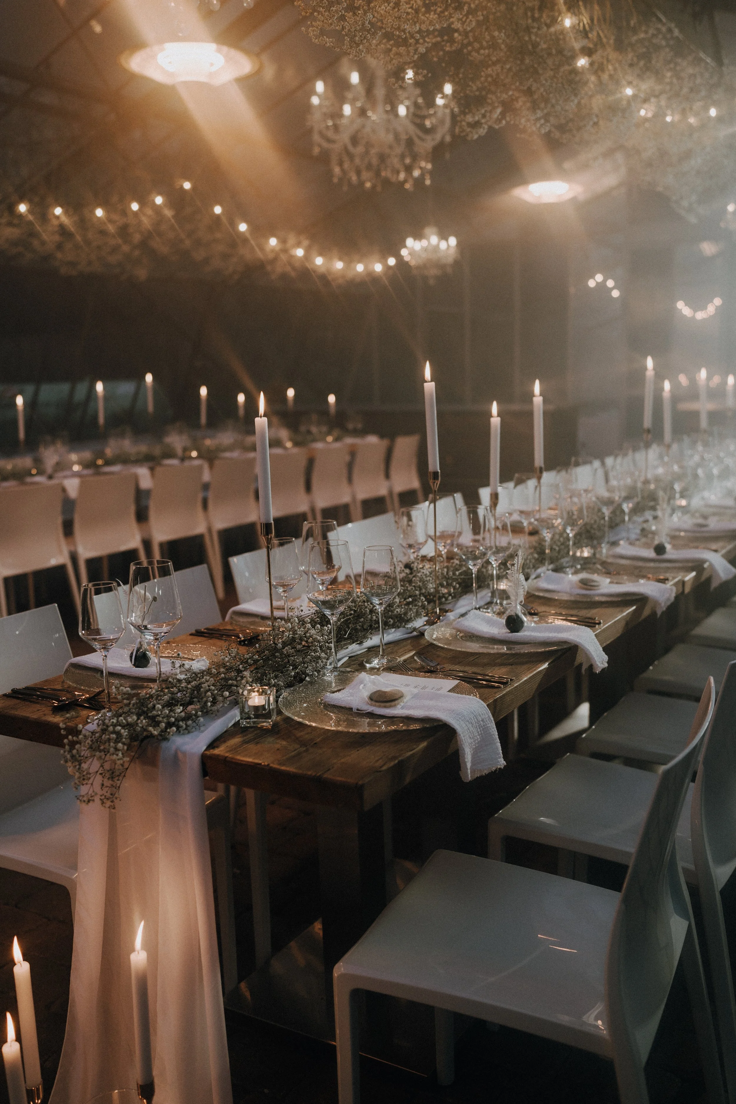 Ein festlich gedeckter Tisch mit Kerzen, Gläsern und Dekoration in einem schlecht beleuchteten, eleganten Raum mit Kronleuchtern und Lichterketten.