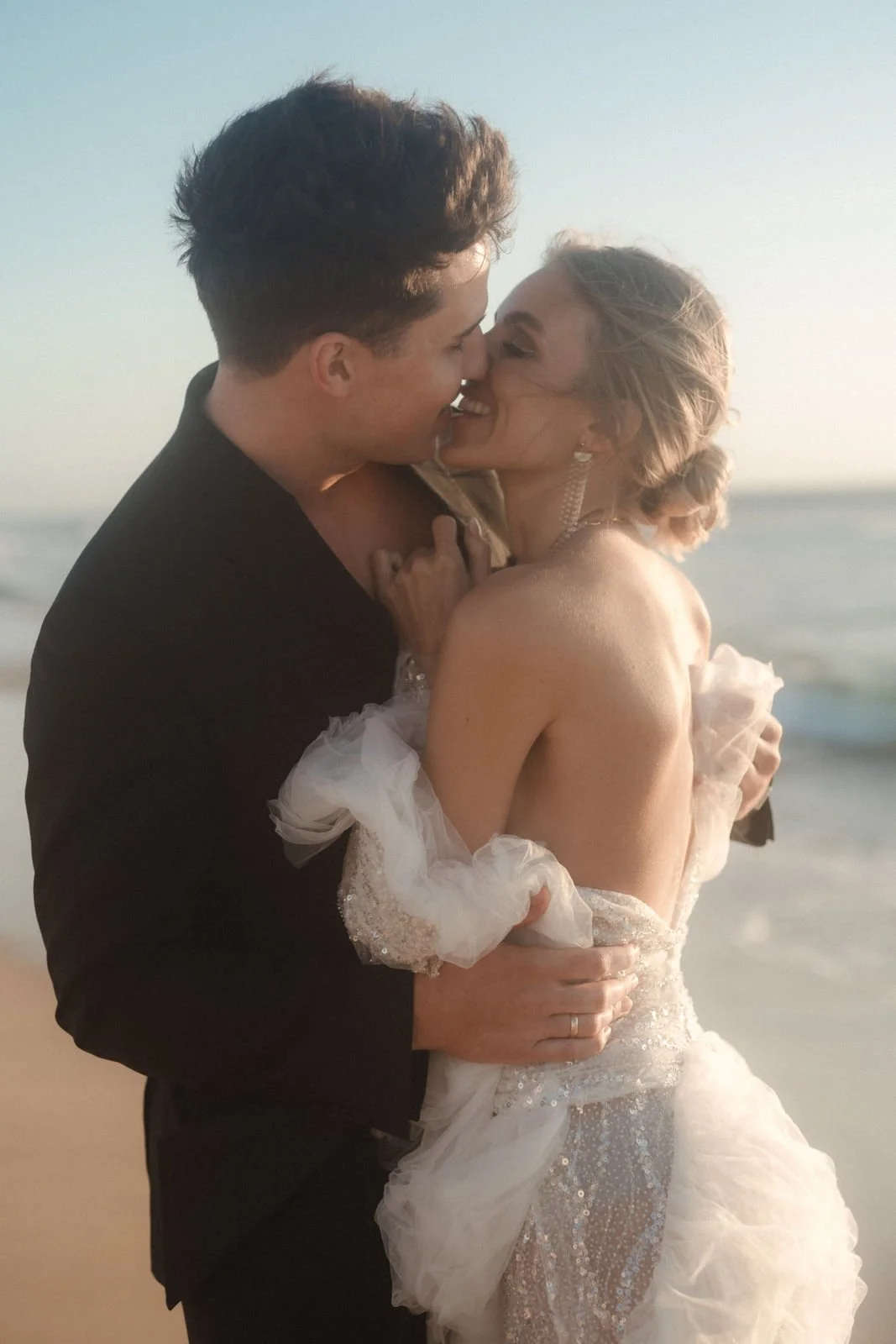 Ein Paar küsst sich an einem Strand bei Sonnenuntergang, offensichtlich an ihrer Hochzeit, mit dem Meer im Hintergrund.