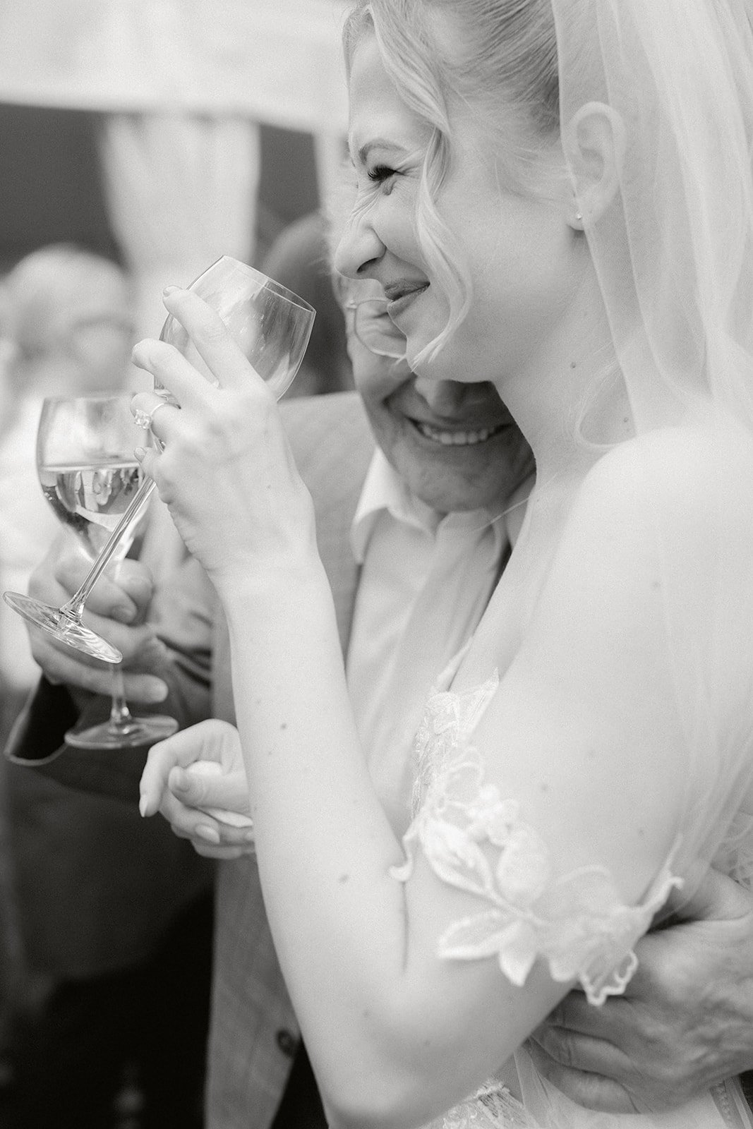 Braut, die auf einer Hochzeit mit einem Glas Wein anstößt, lächelnd und umarmt von einem Mann.