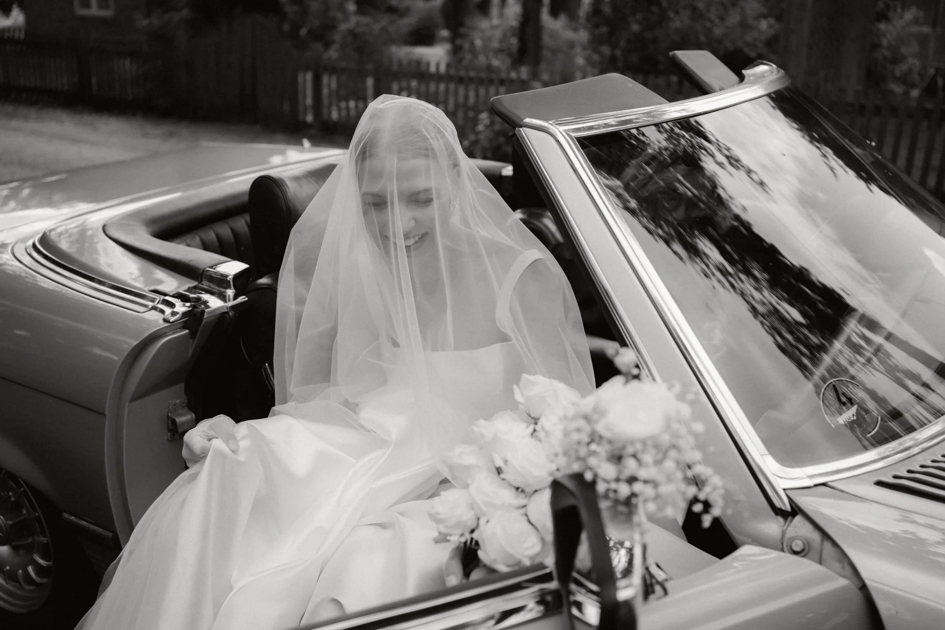 Braut in Hochzeitskleid und Schleier sitzt in einem Cabriolet, lächelt, mit Blumen im Vordergrund, im Freien, schwarz-weiß Fotografie.