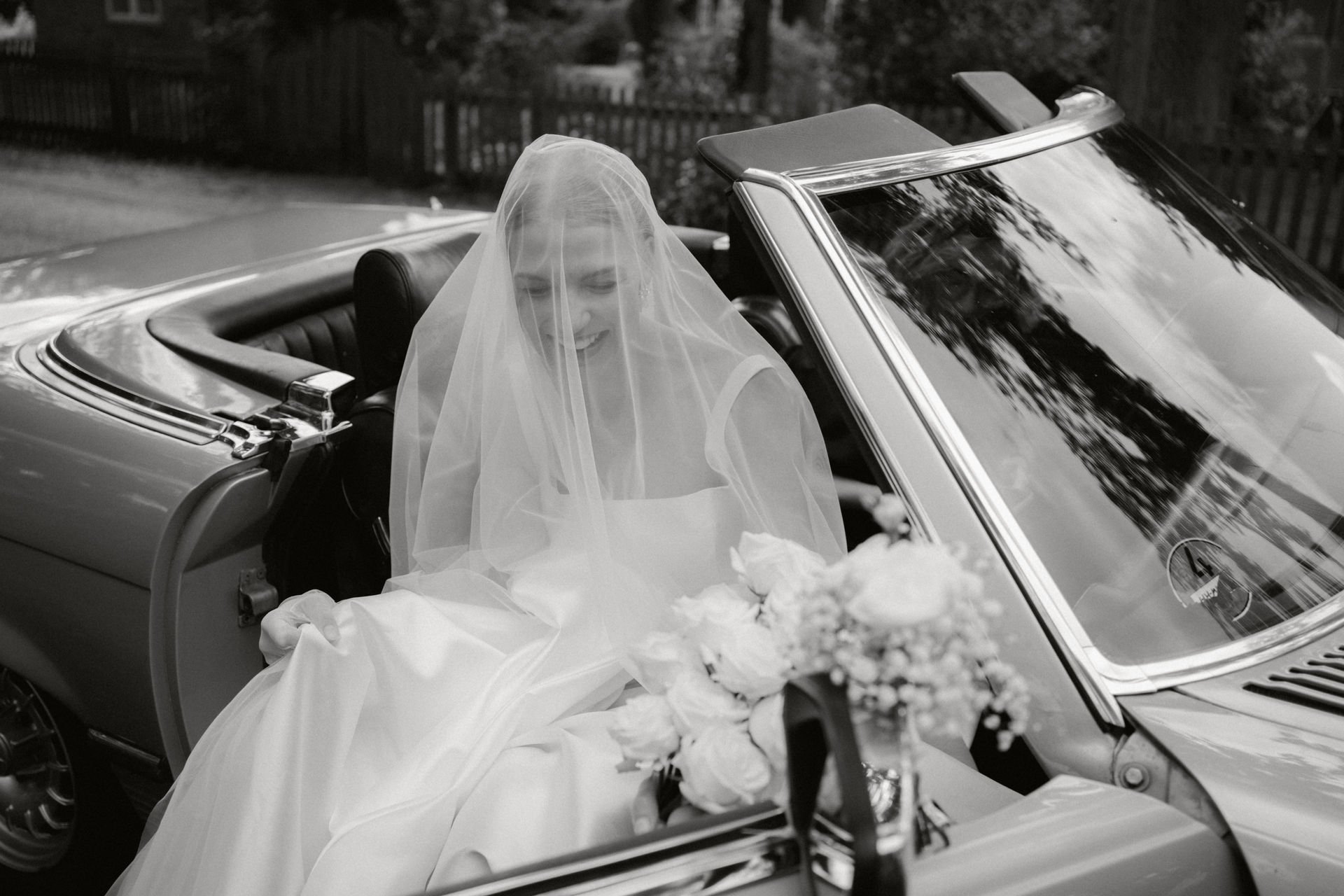 Braut in Hochzeitskleid und Schleier sitzt in einem Vintage-Stretch-Limousine mit offenem Verdeck, Hintergrund aus Bäumen und Zaun, Schwarz-Weiß-Fotografie.