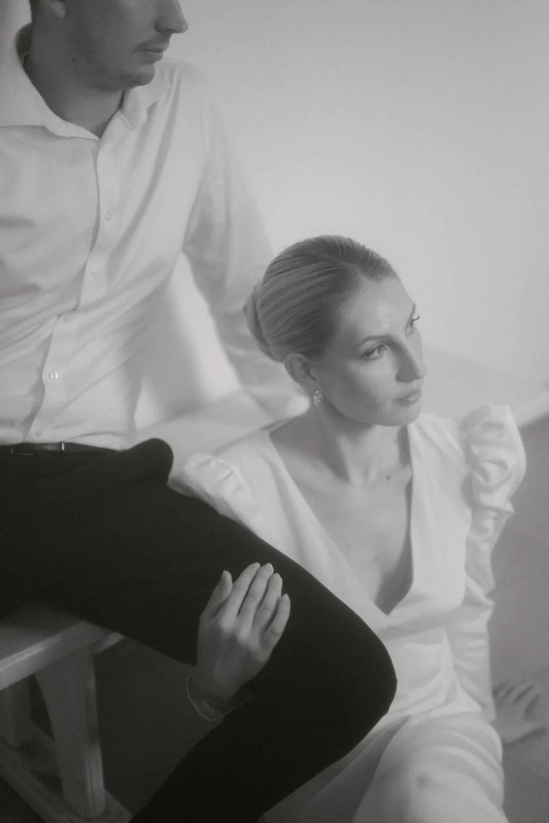 Schwarz-Weiß-Foto eines Mannes im weißen Hemd, der neben einer sitzenden Frau mit hochgestecktem Haar und weißen Kleid sitzt, die nachdenklich blickt.