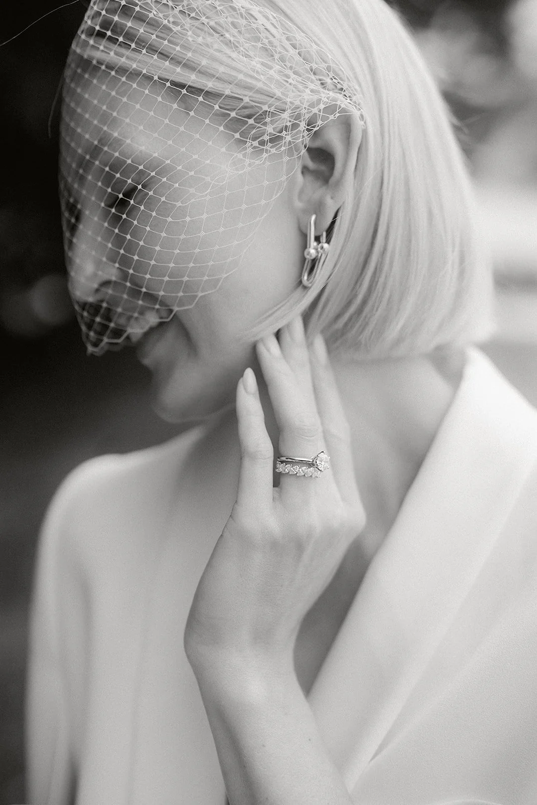 Schwarz-weiß-Fotografie einer Frau mit einem Schleier über dem Gesicht, tragende eleganten Schmuck, inklusive Ring und Ohrringen, die an einer Hochzeit oder einem formellen Anlass erinnert.