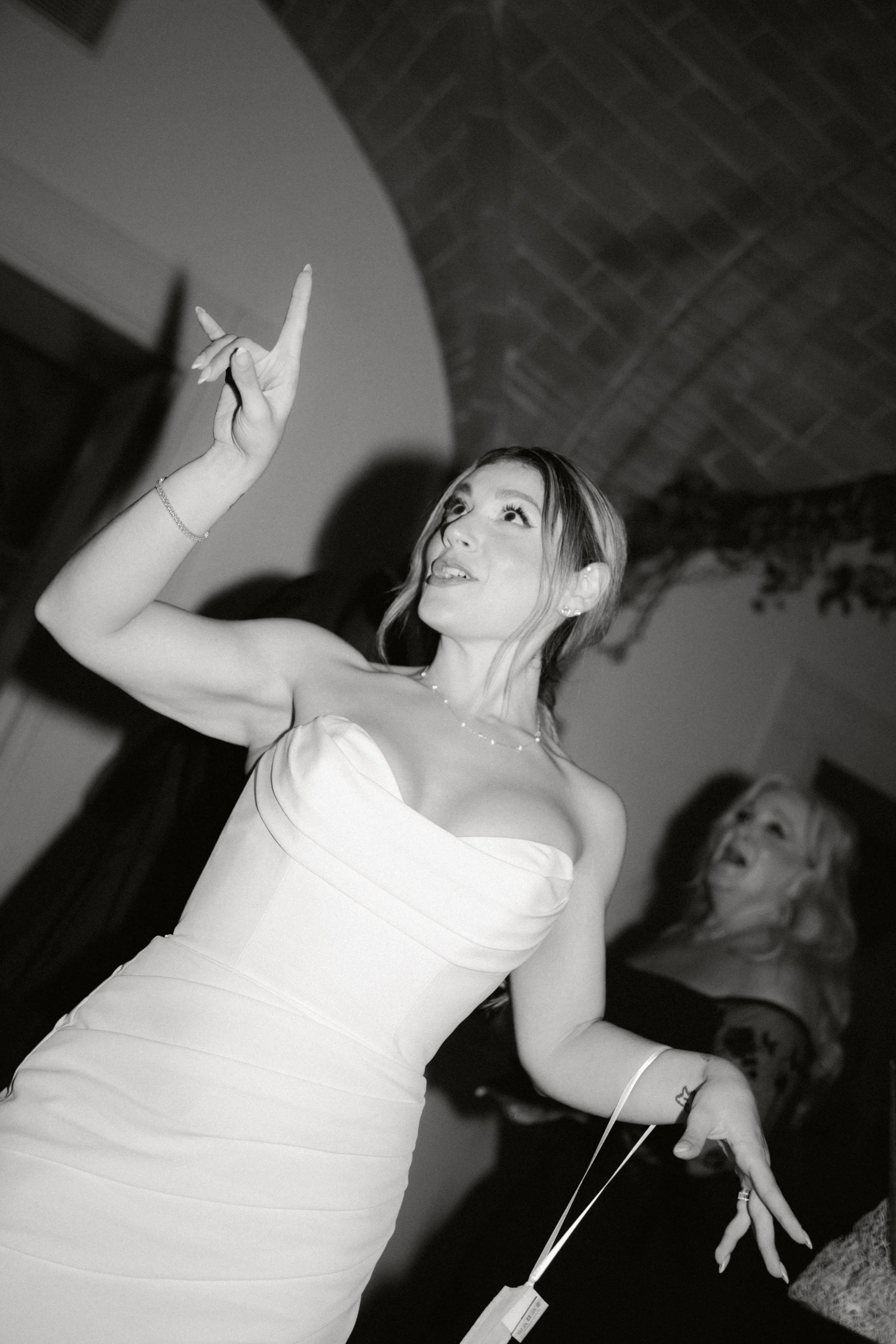 Eine lachende Frau in einem weißen Kleid, die tanzt und ihre Arme hebt, auf einer Party im Innenraum.