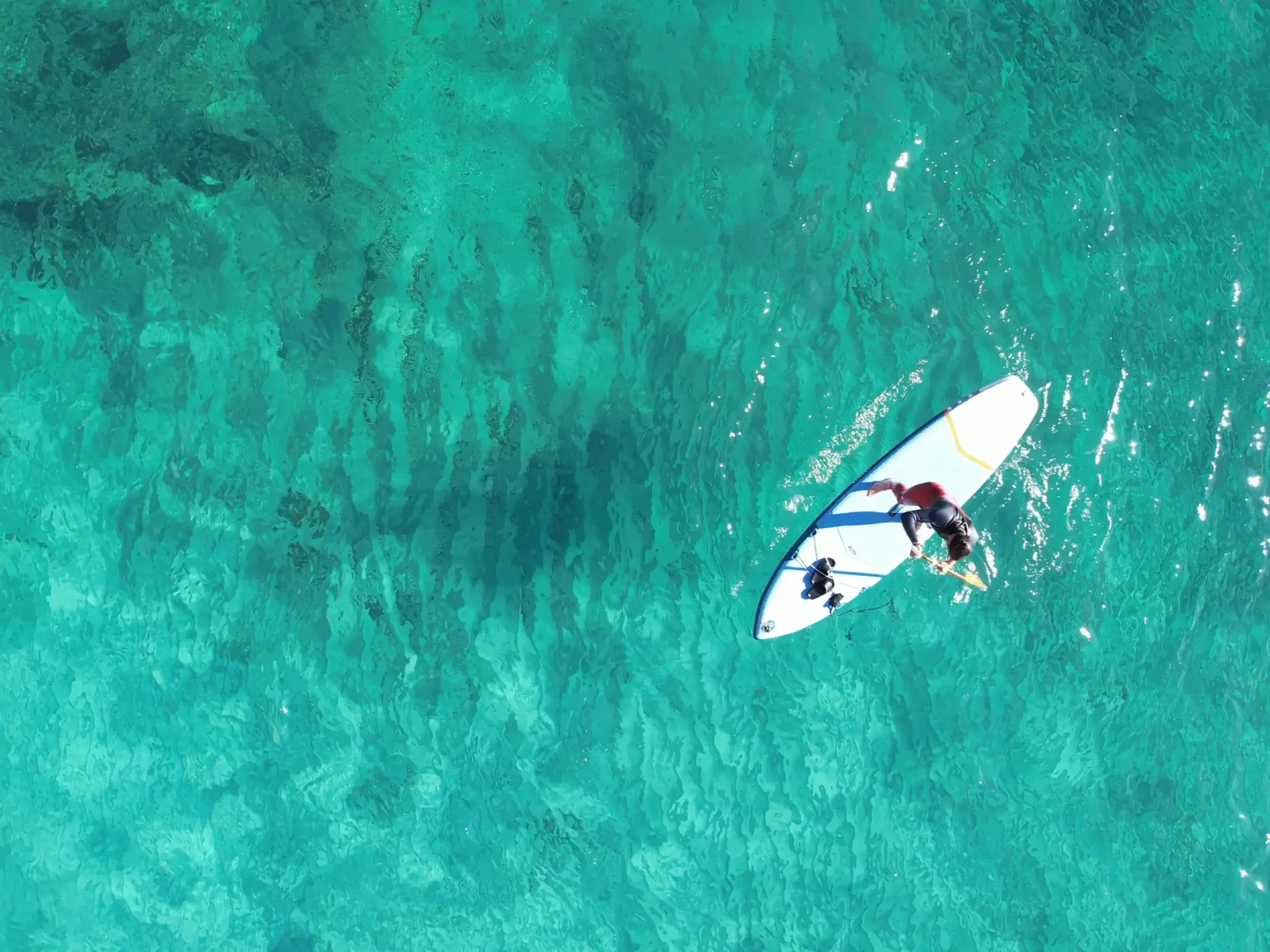 An aerial view of a person stand-up paddleboarding on clear turquoise water.