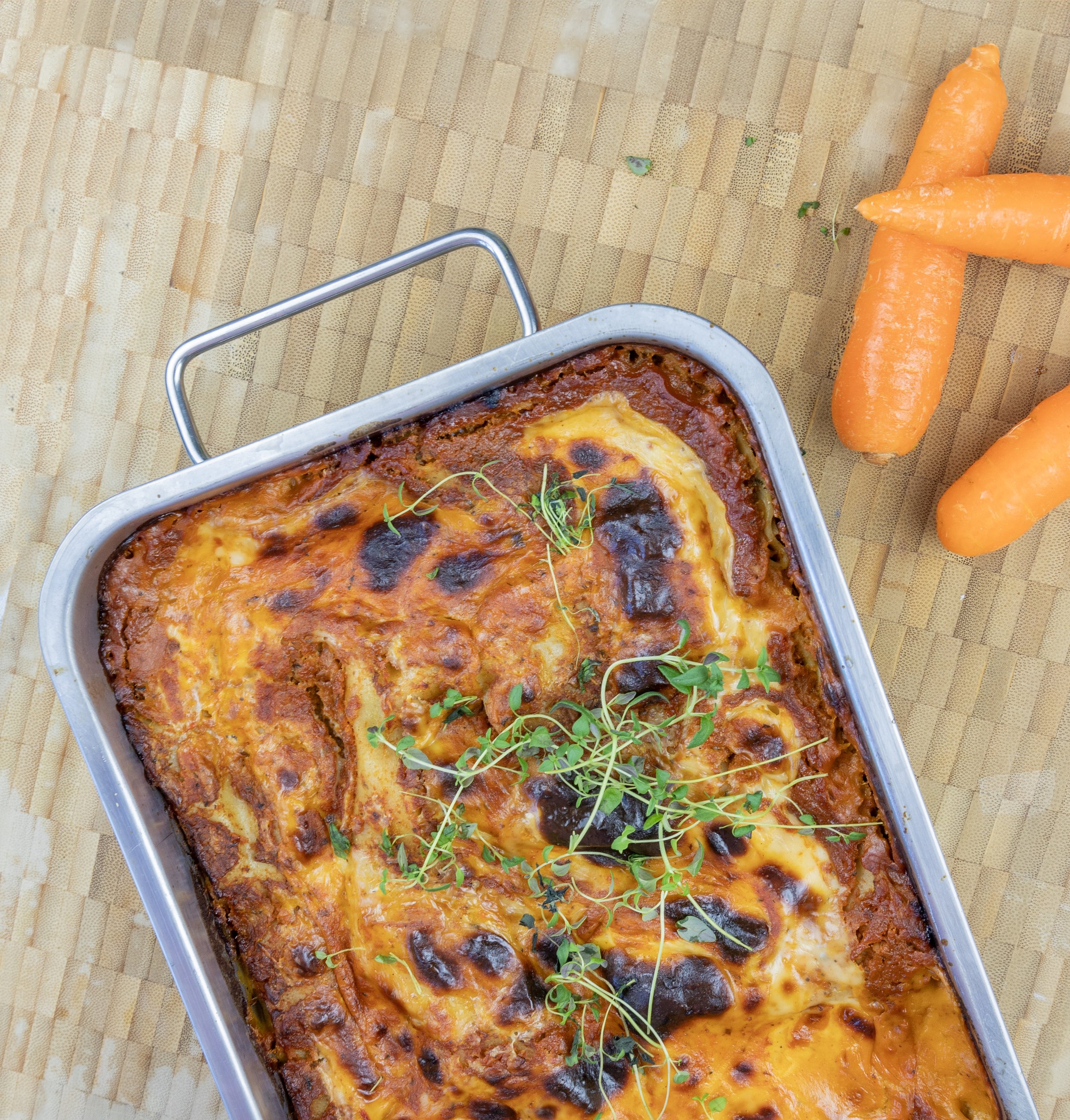 Lasagne med ost, tomatsauce og friske urter i en ovnfast fad, med gulerødder ved siden på et træbord.