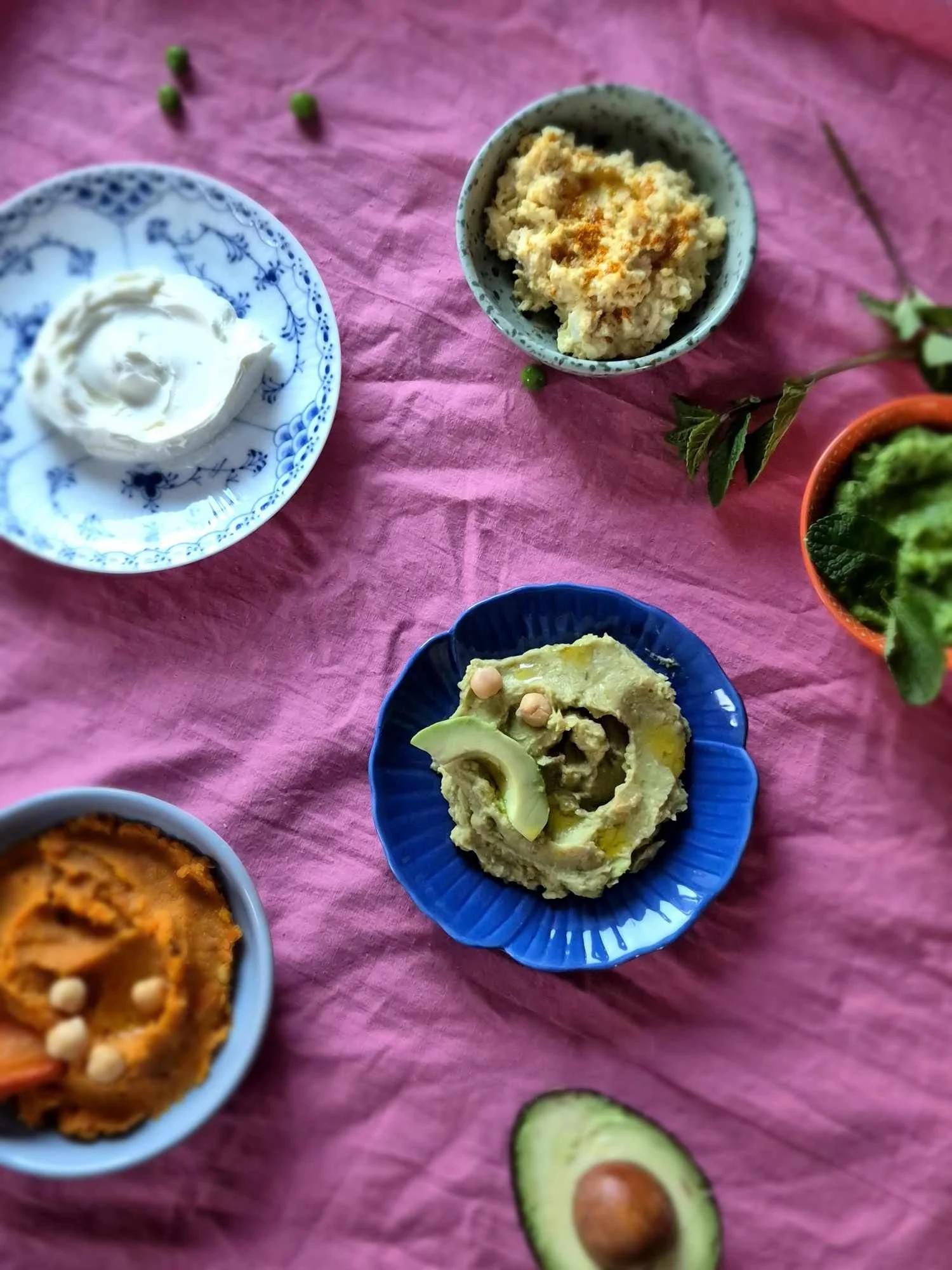 Forskellige retter og dip sauces på farvetak på en pink klud, inklusive guacamole, hummus, en creme og en smøragtig dip, med avocado, agurk og grønne ærter.