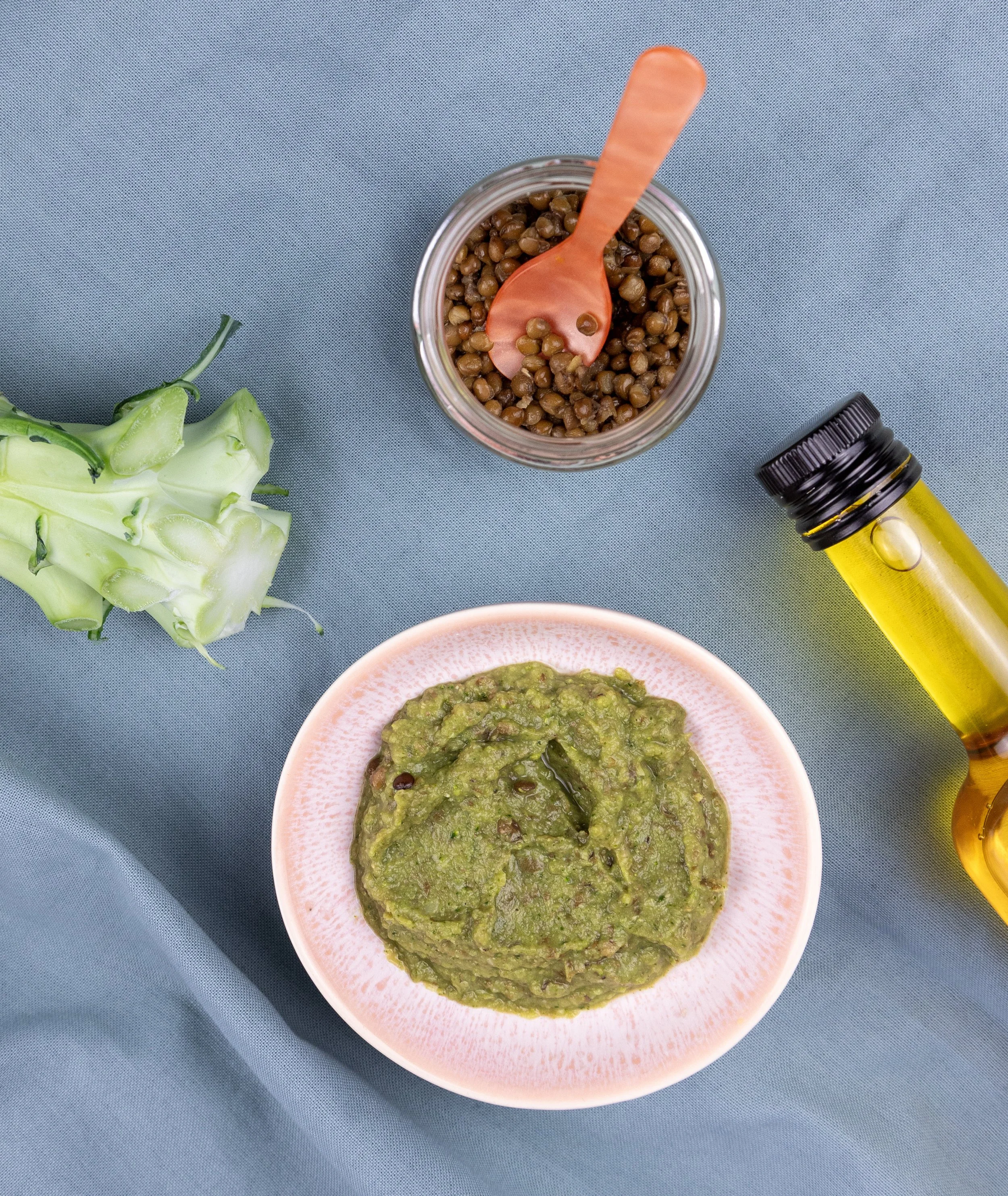 En skål med guacamole, en glasboks med brune linser, en flaske med olie, og en grøn grøntsag på en blå baggrund.