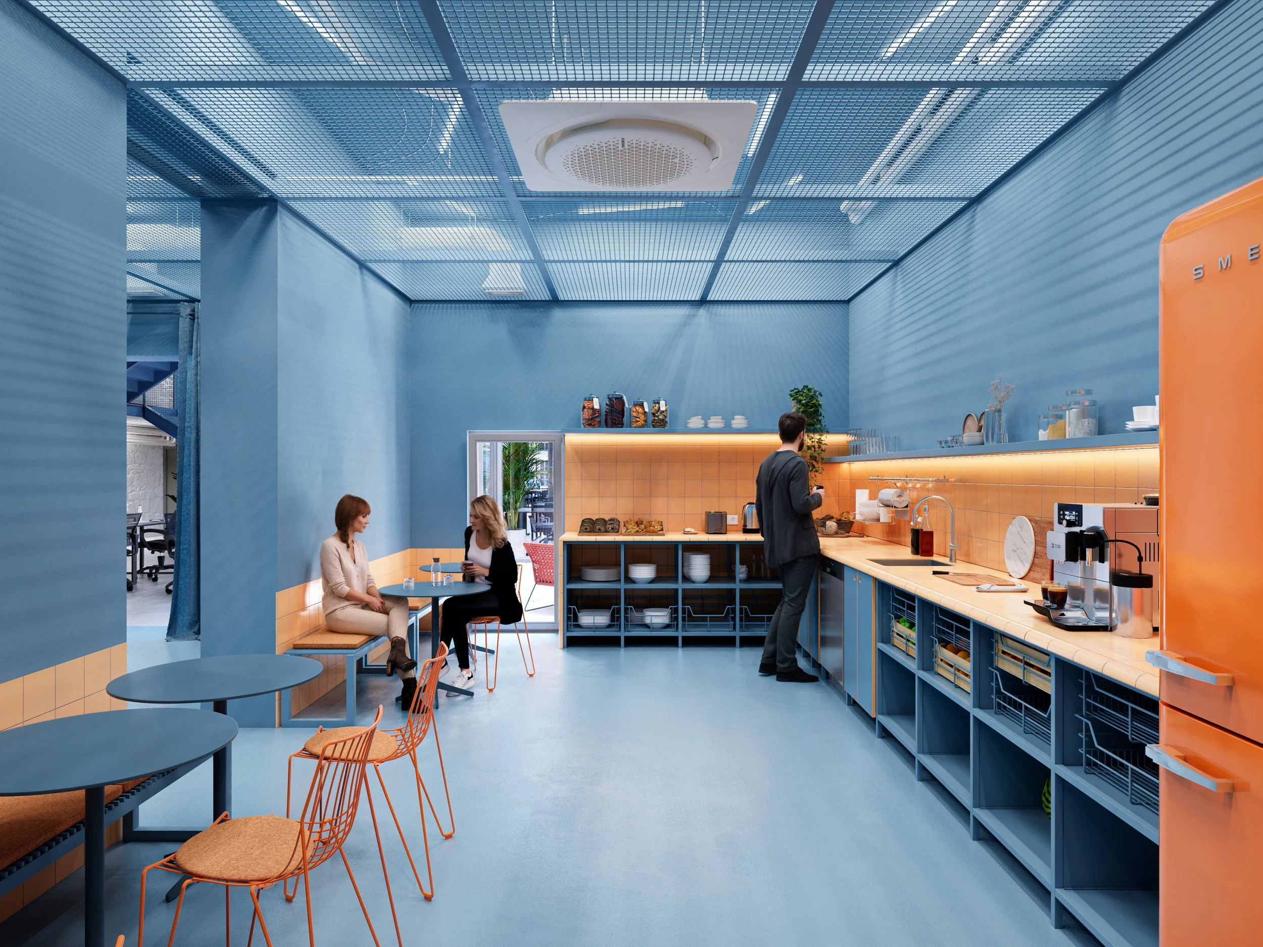 Modern cafeteria with blue and orange decor, two women seated at a small table, a man preparing coffee at a counter, and bright lighting.