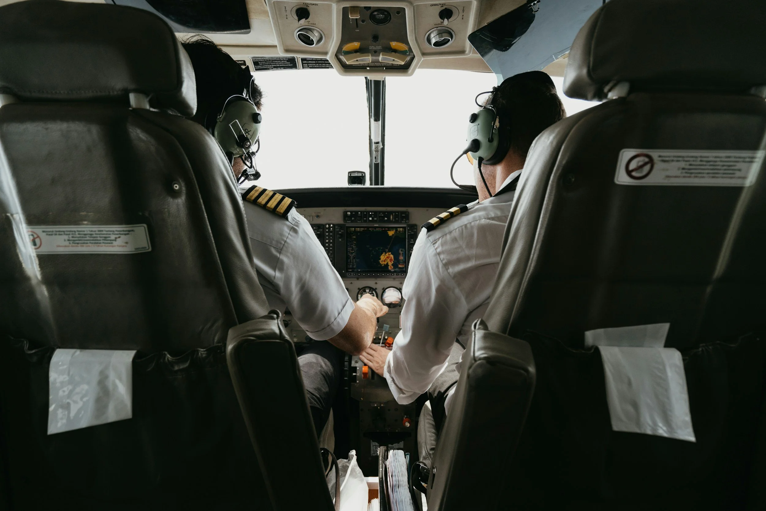 Two pilots in a cockpit flying an aircraft, both wearing headsets and white uniforms with shoulder epaulets.