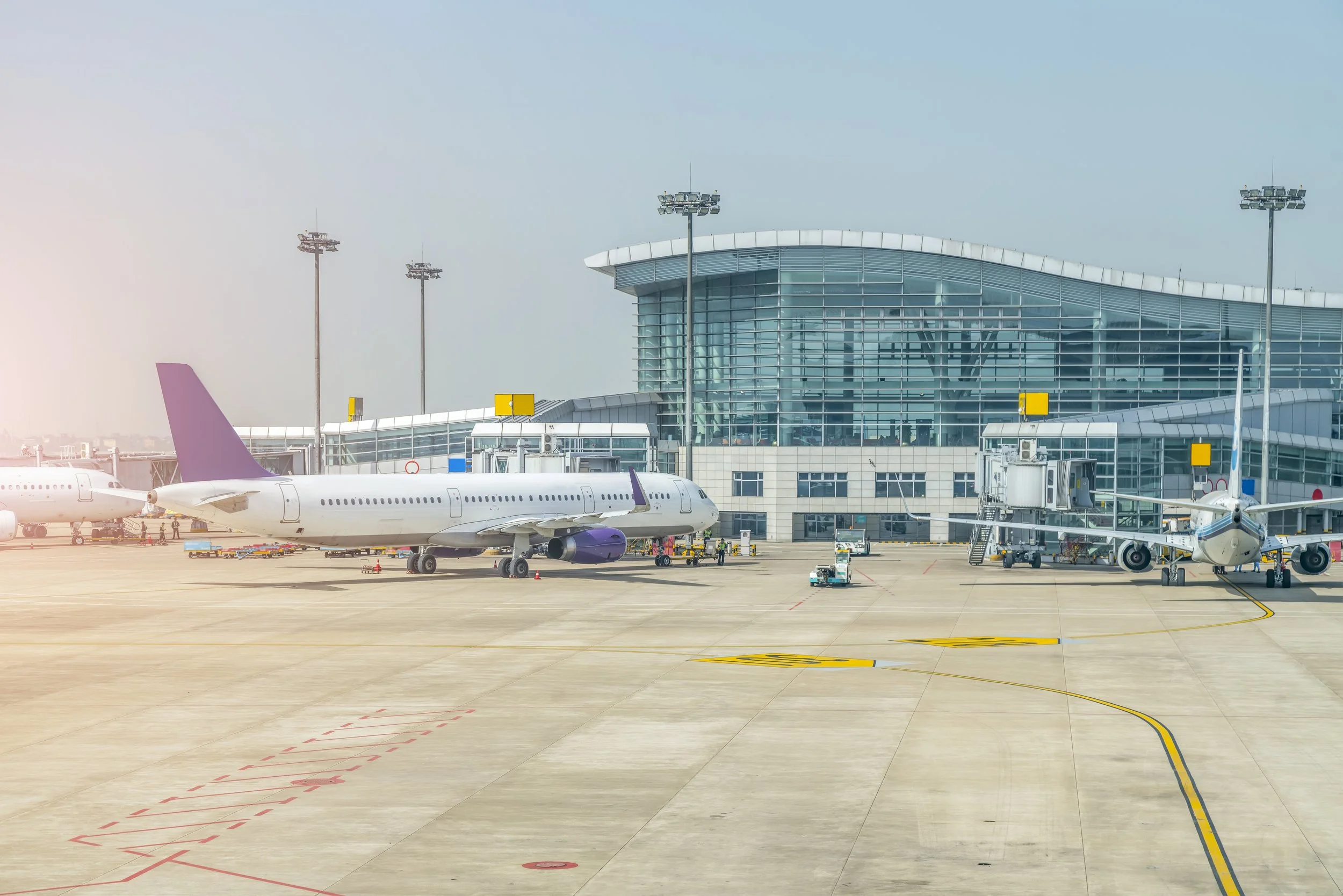 Airport terminal building with aircraft parked at gates, jet bridges connected to planes, and ground vehicles nearby.