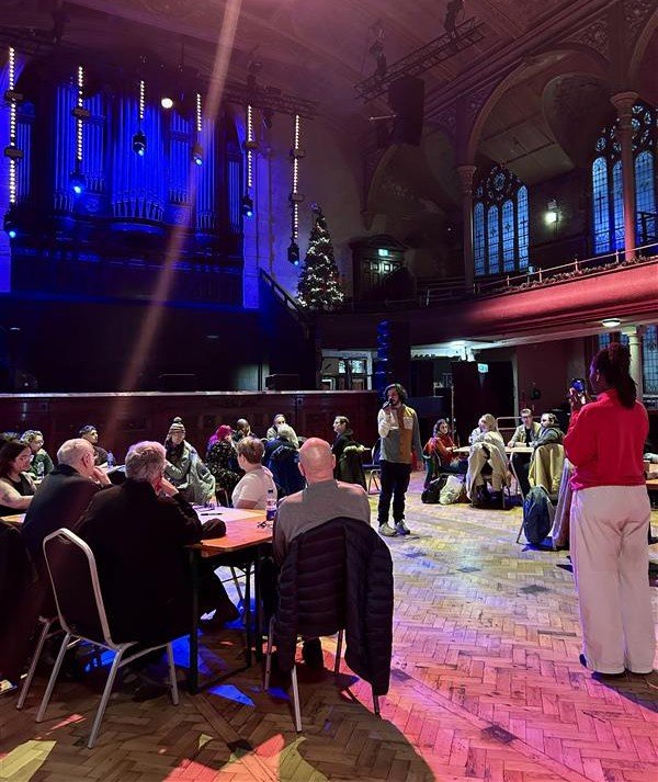 People sit around tables inside the Albert Hall in Manchester during a live discussion event, with stage lighting, a large organ, and stained-glass windows in the background.
