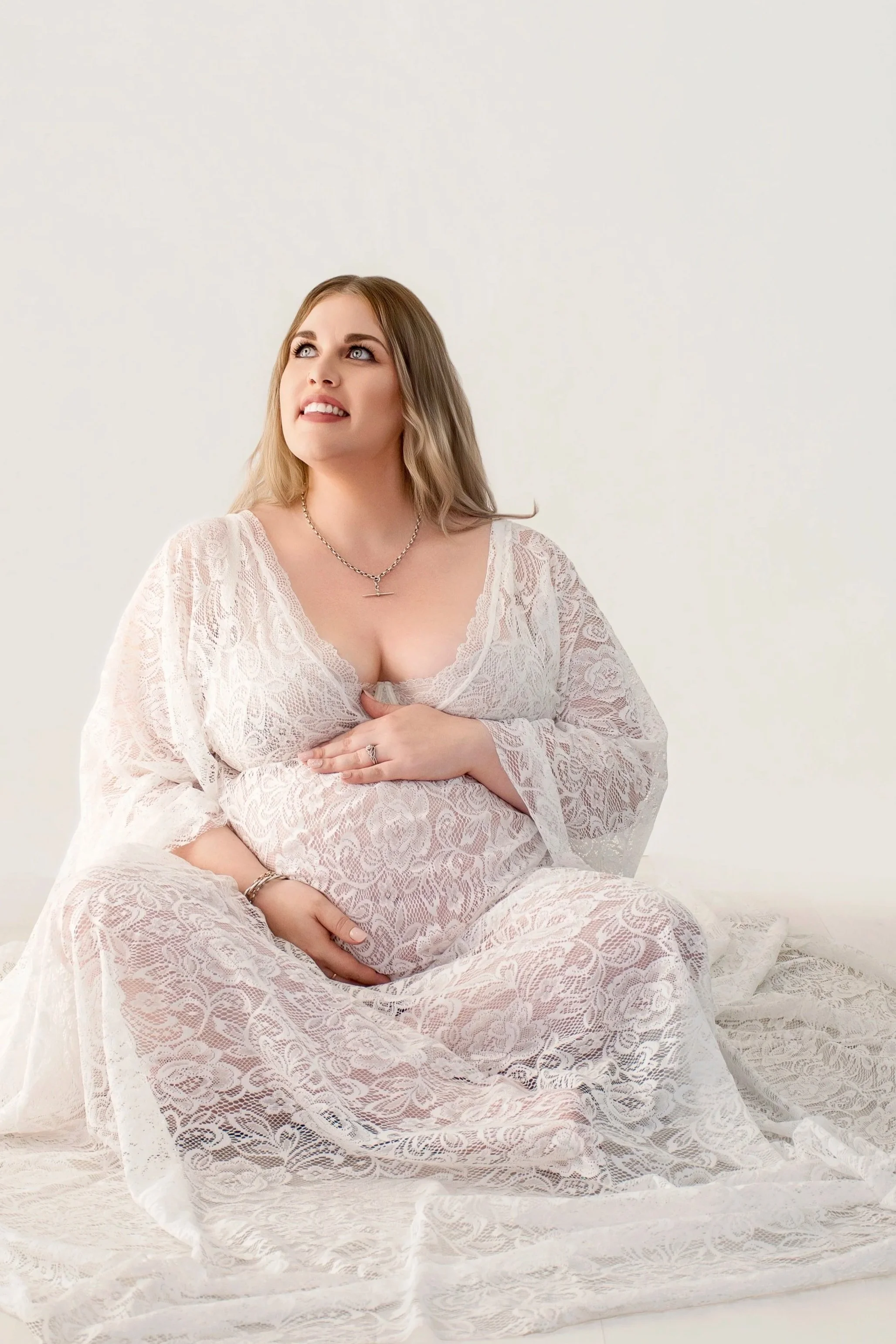 A pregnant woman in a white lace dress, sitting on a lace-covered surface, looking up with a thoughtful expression.