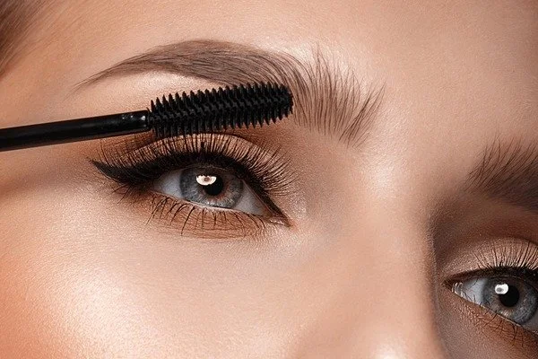Close-up of a woman's face applying mascara to her eyebrow with a mascara wand.