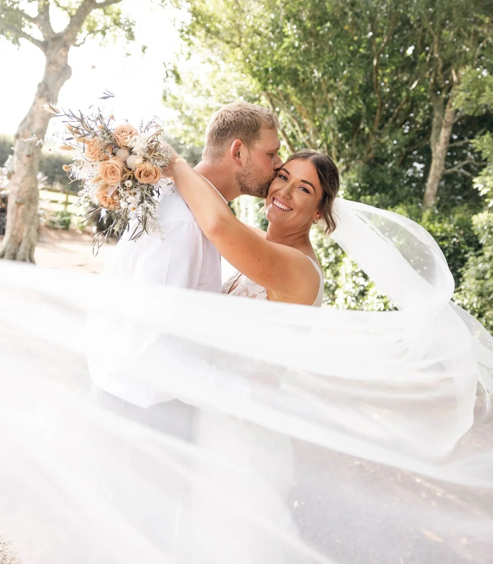 A newlywed couple embracing outdoors, with the groom kissing the bride on the cheek, both smiling. The bride is holding a bouquet of flowers, and her veil is flowing around them. Green trees are in the background.