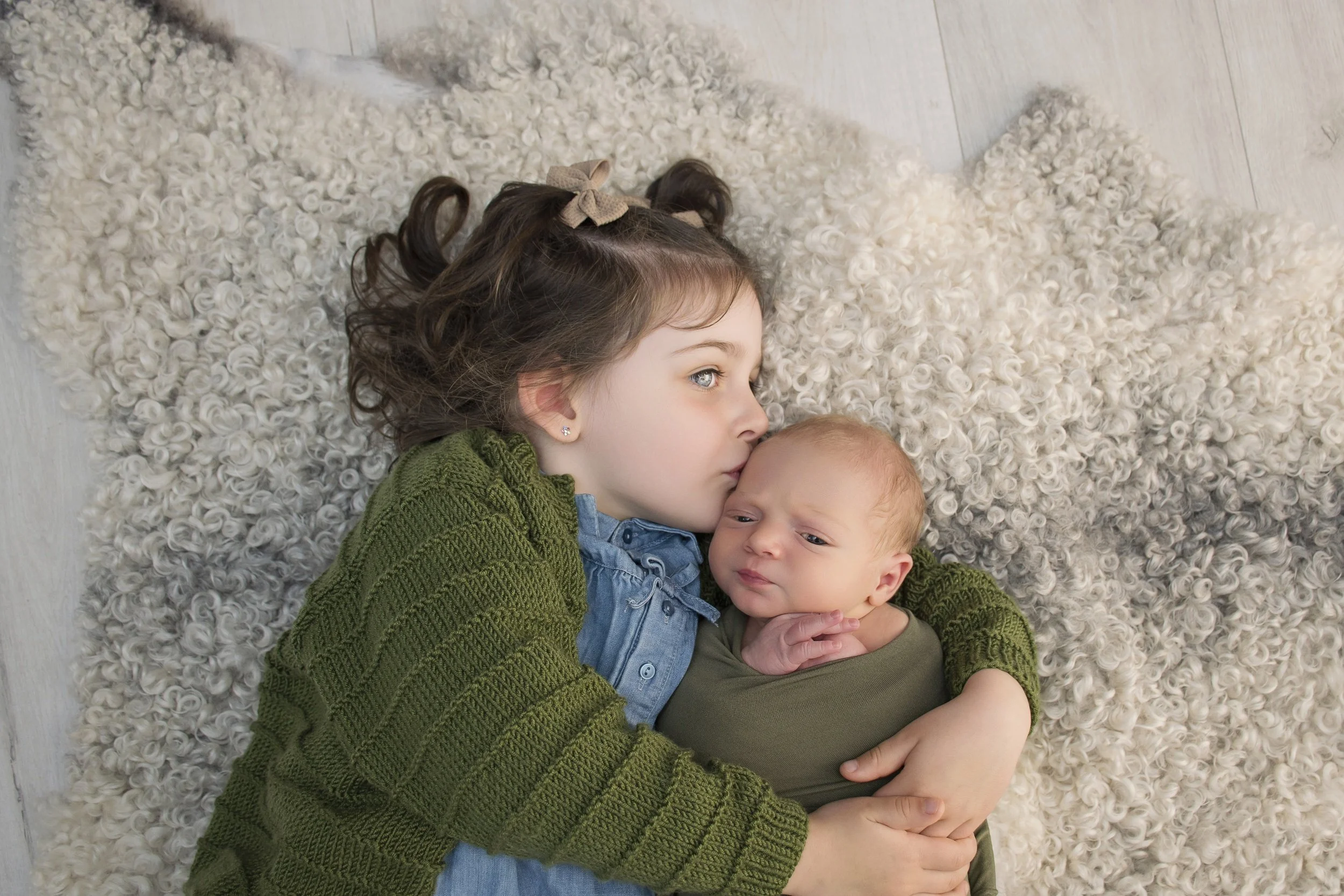A young girl with curly brown hair and blue eyes lying on a fluffy white rug, holding and kissing a newborn baby with light brown hair and blue eyes.