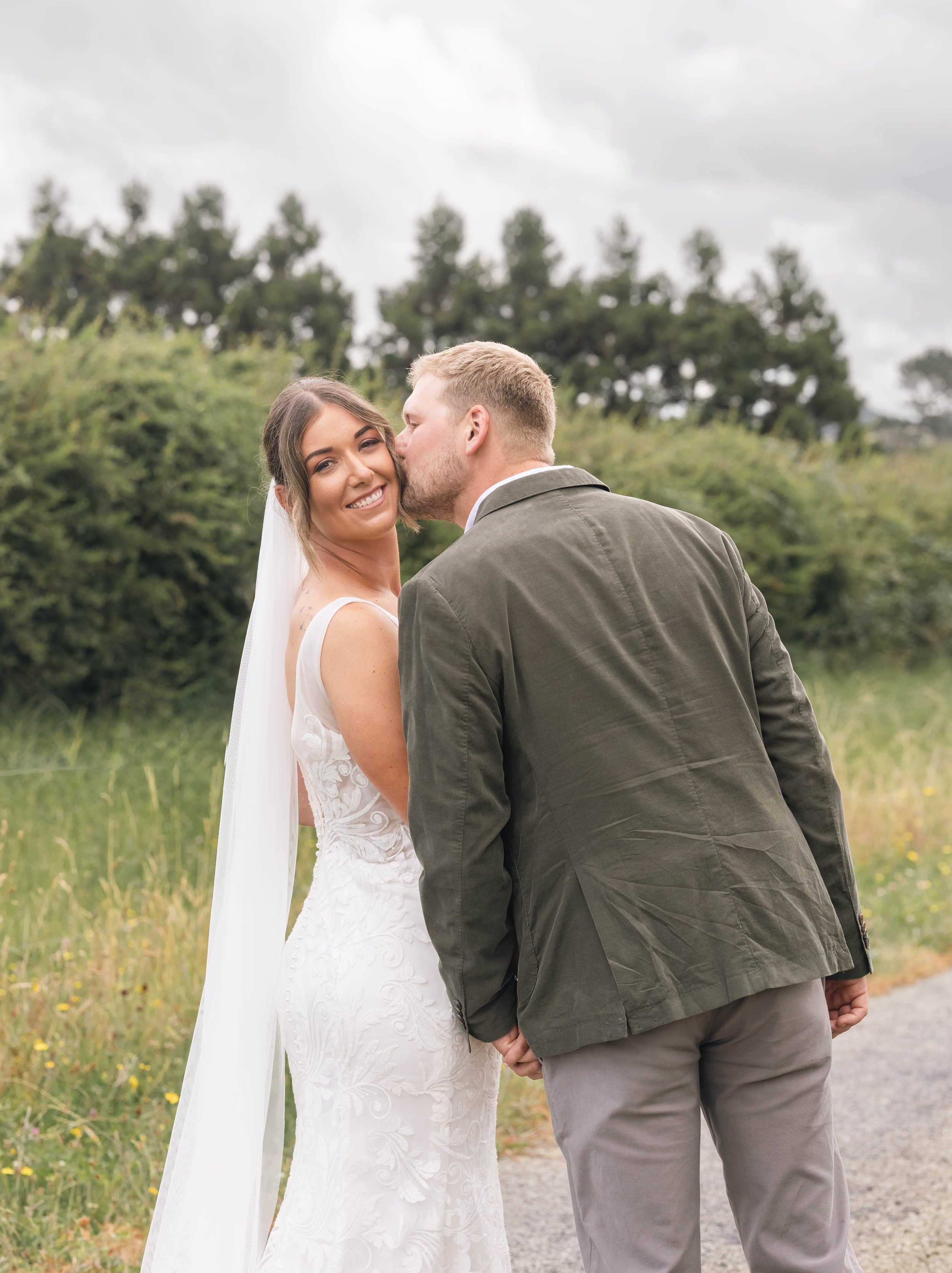 A bride and groom outdoor wedding photo, groom kissing bride cheek, bride smiling, trees and cloudy sky in background.