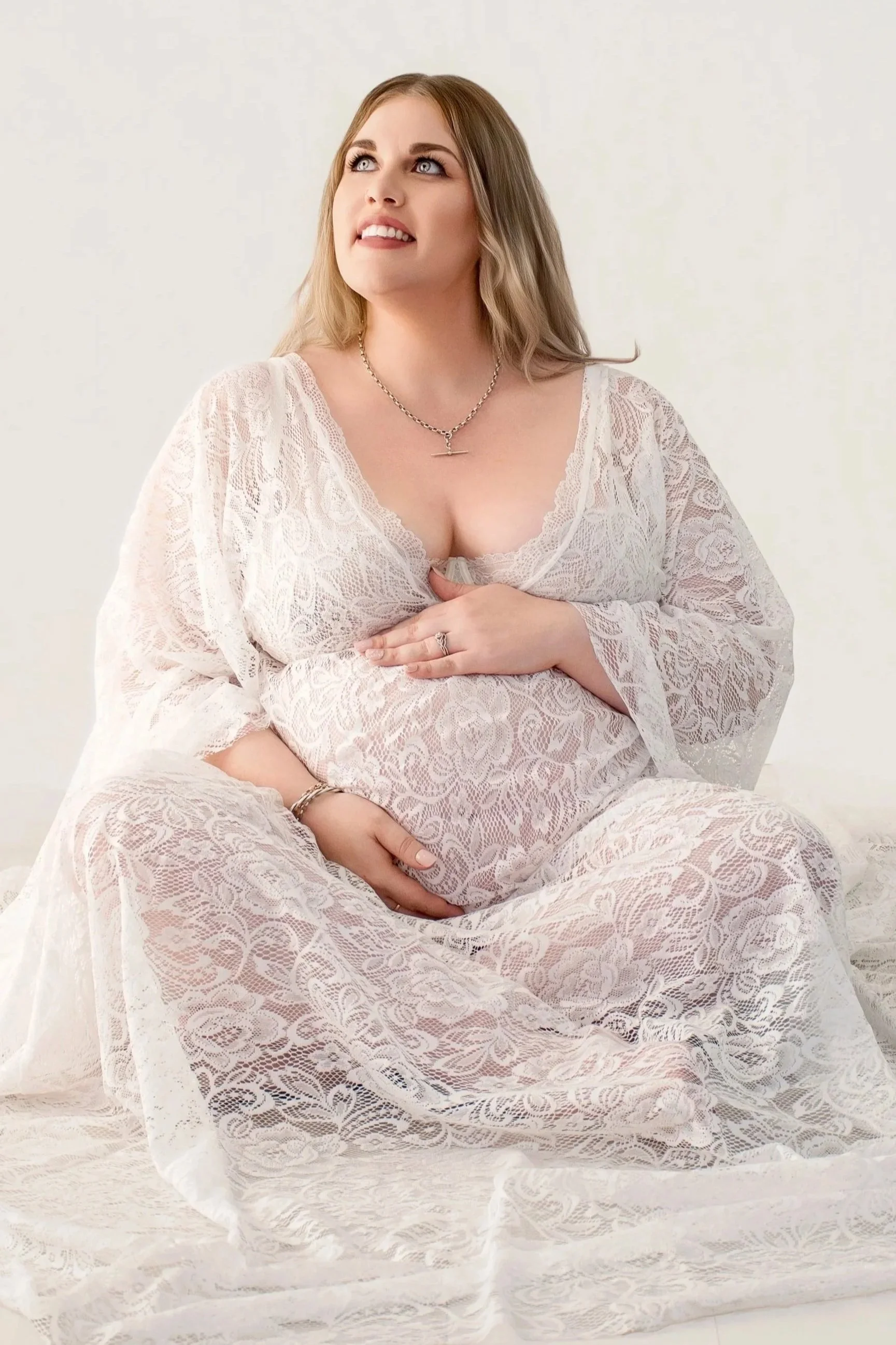 A pregnant woman sitting on the floor, wearing a white lace dress, with her hands on her belly, looking upward.