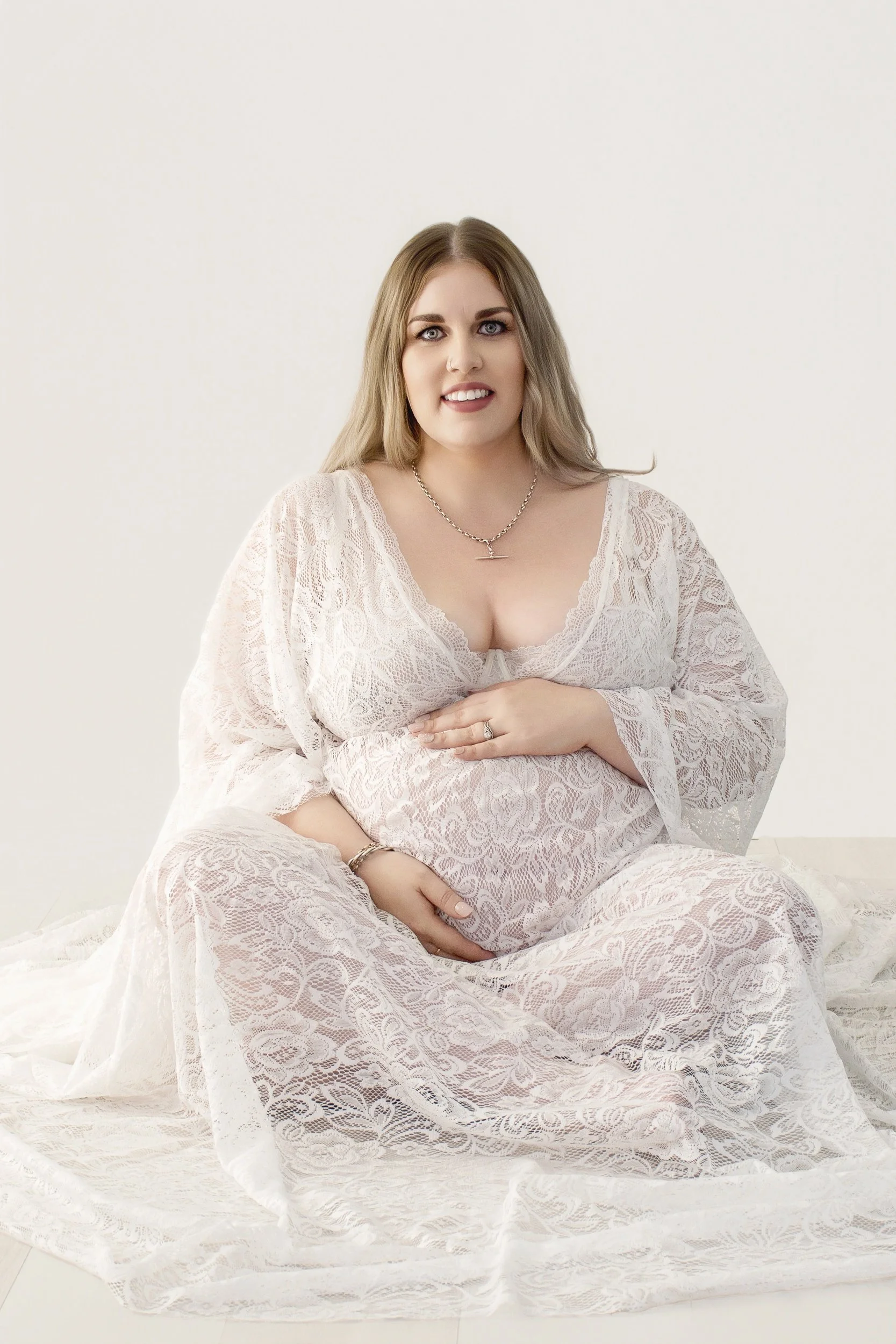 A pregnant woman sitting on the floor wearing a white lace dress, holding her belly with one hand, smiling, with a white background.