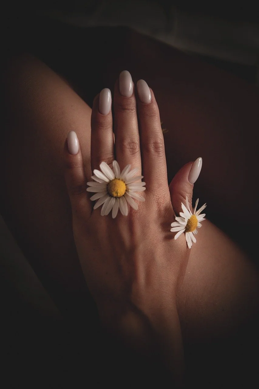 A hand with manicured nails and daisy flowers on two fingers, against a dark background.