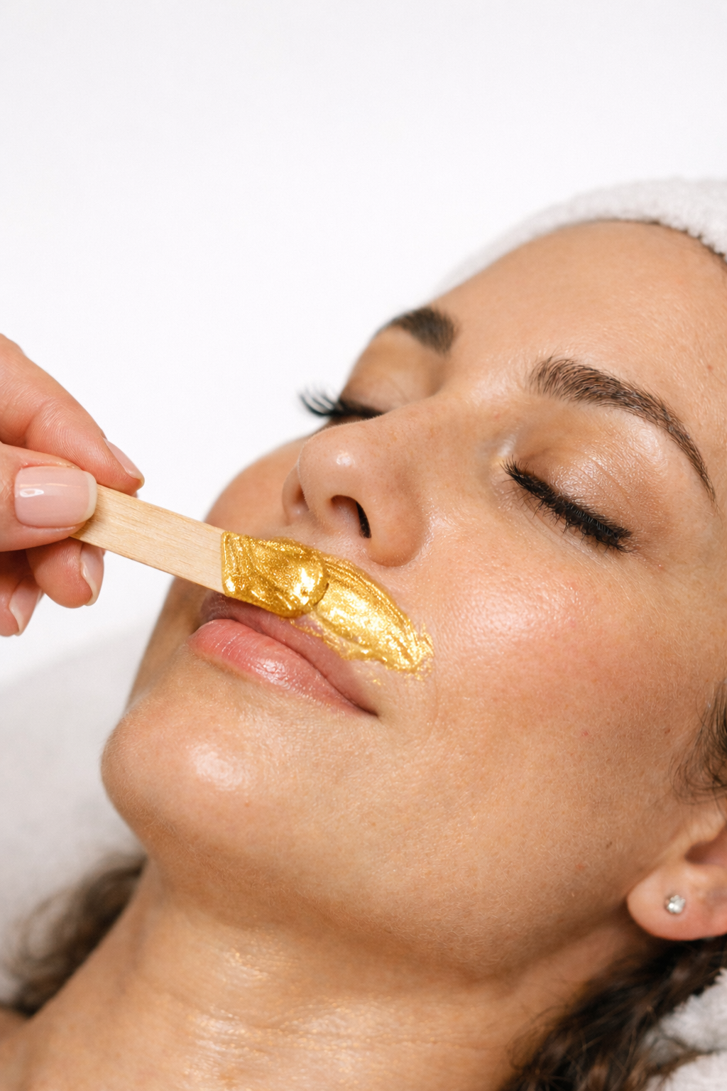 A woman with closed eyes receiving a facial treatment, applying a gold-colored facial wax with a spatula for smooth upper lip
