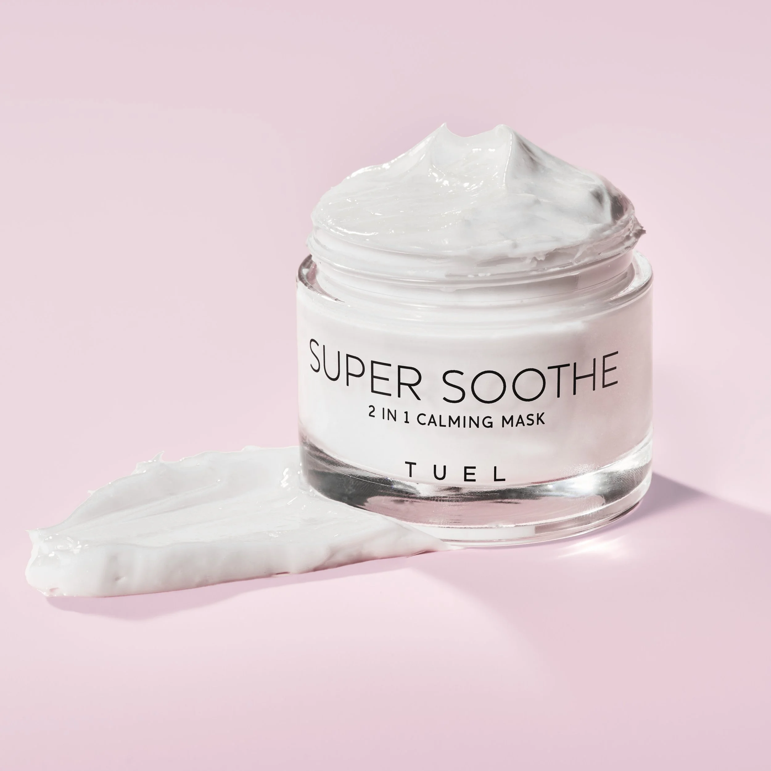 A jar of Super Soothe 2-in-1 calming mask with some of the white cream spread on a pink surface.