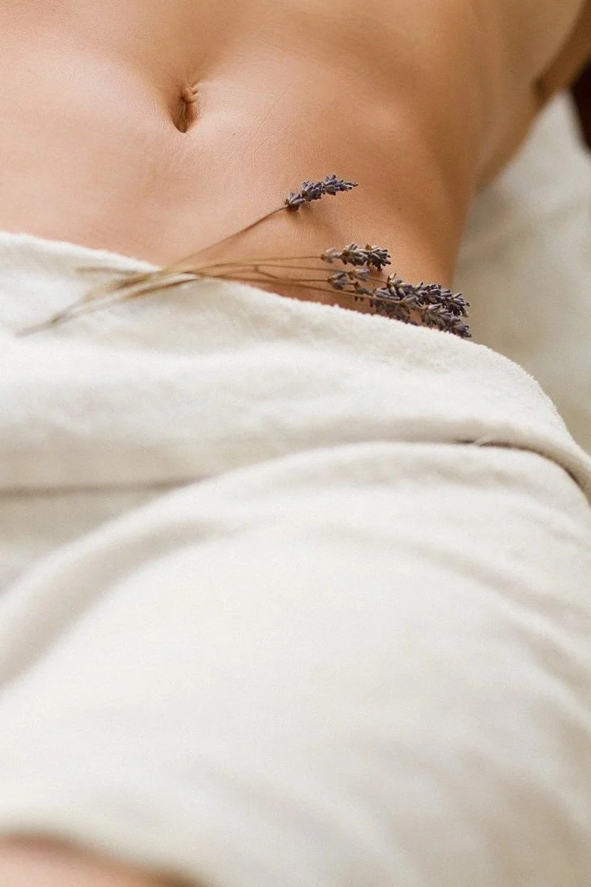 Close-up of a person's bare abdomen with a small sprig of lavender resting on their stomach, partially covered by a light-colored blanket or sheet.