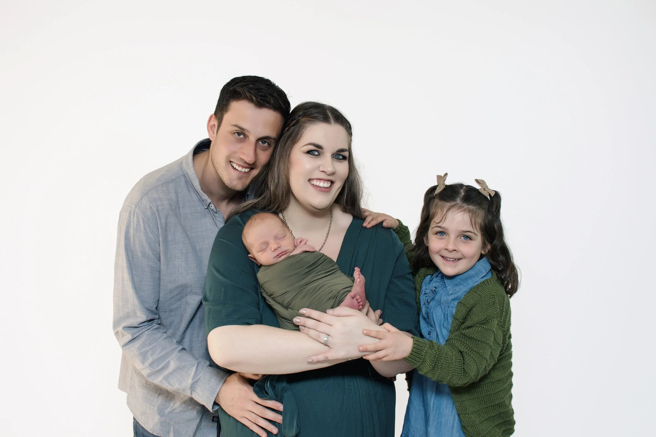 A happy family of four, including a mother holding a sleeping baby, a father, and a young girl smiling, standing against a plain white background.