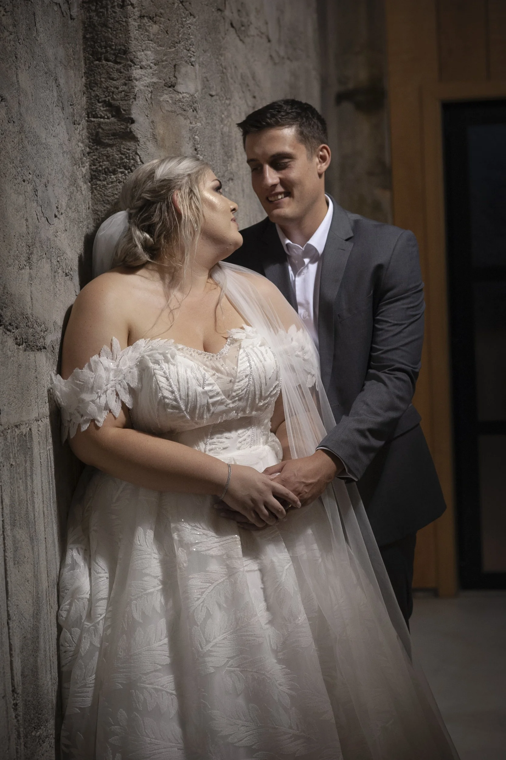 A bride and groom in wedding attire sharing an intimate moment, with the bride leaning against a concrete wall and the groom holding her hands, inside a rustic indoor setting.