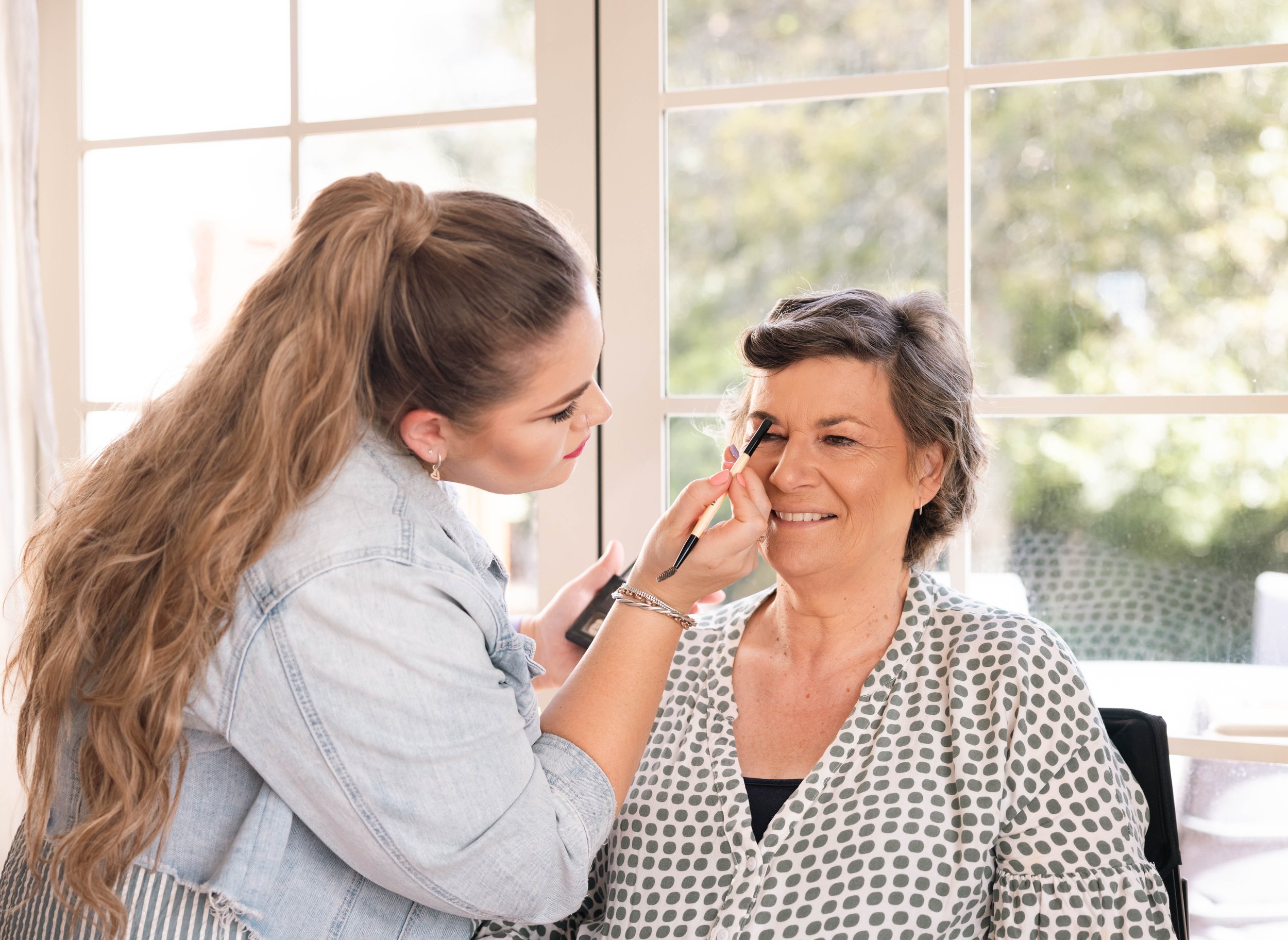 A makeup artist applying makeup to a smiling older woman sitting by a large window.