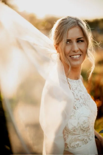 A smiling woman in a white lace dress holding a flowing white fabric outdoors during sunset.