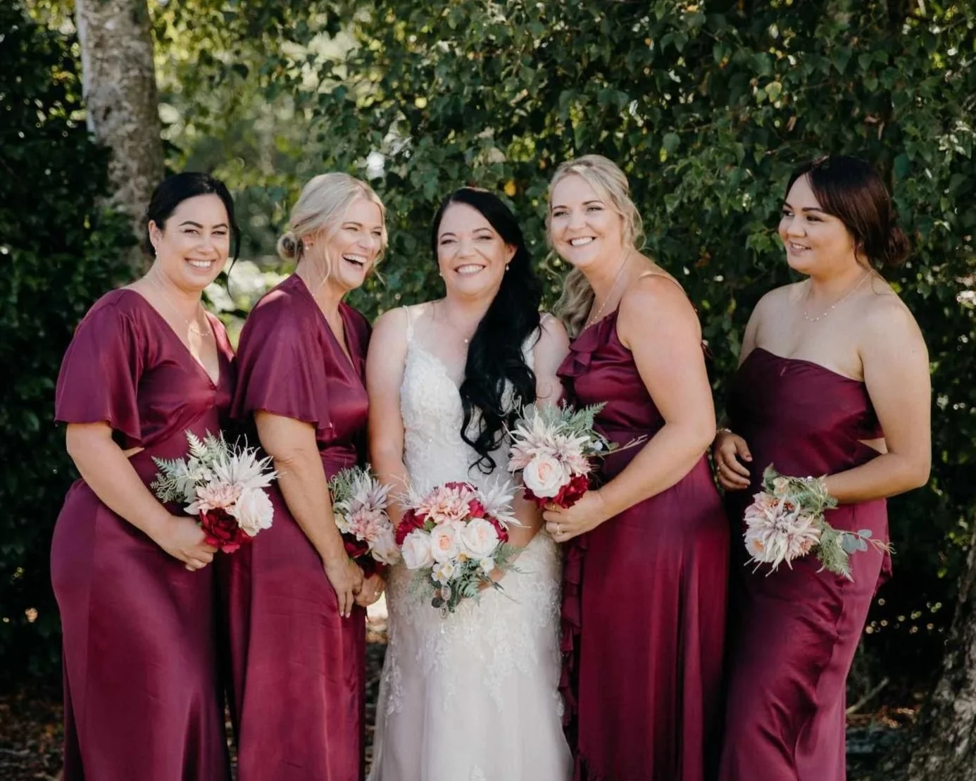 A bride in a white wedding dress standing with four bridesmaids in burgundy dresses, all holding bouquets of flowers, outdoors with trees and greenery in the background.