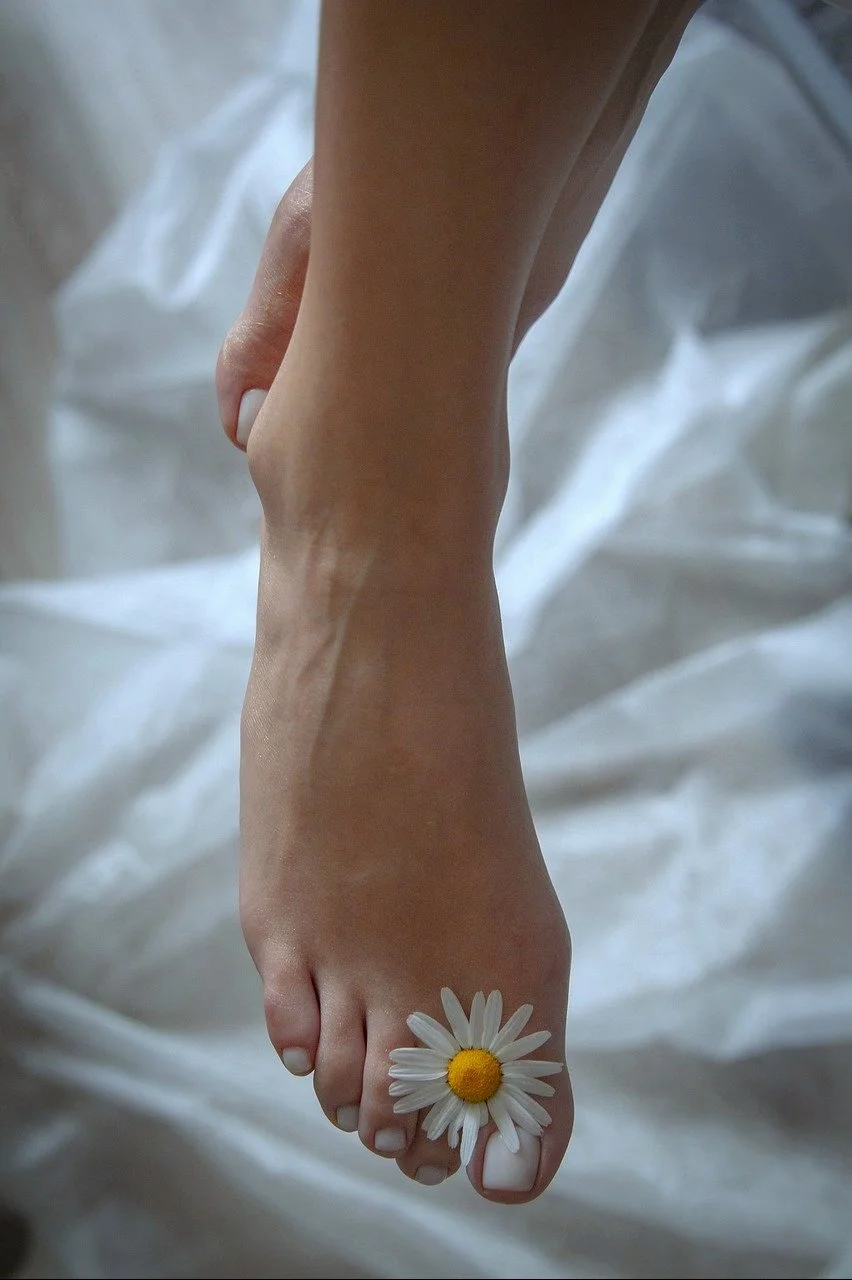 Close-up of a person's foot with a white daisy flower placed on the toes, resting on white fabric.