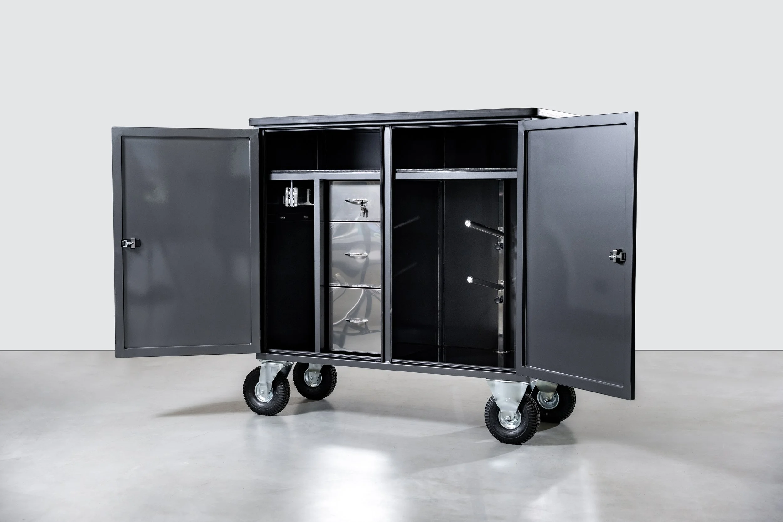 A large metal storage cabinet with open doors on wheels, containing drawers and various tools or equipment, placed outdoors in front of a building with large glass windows and a tiled roof.
