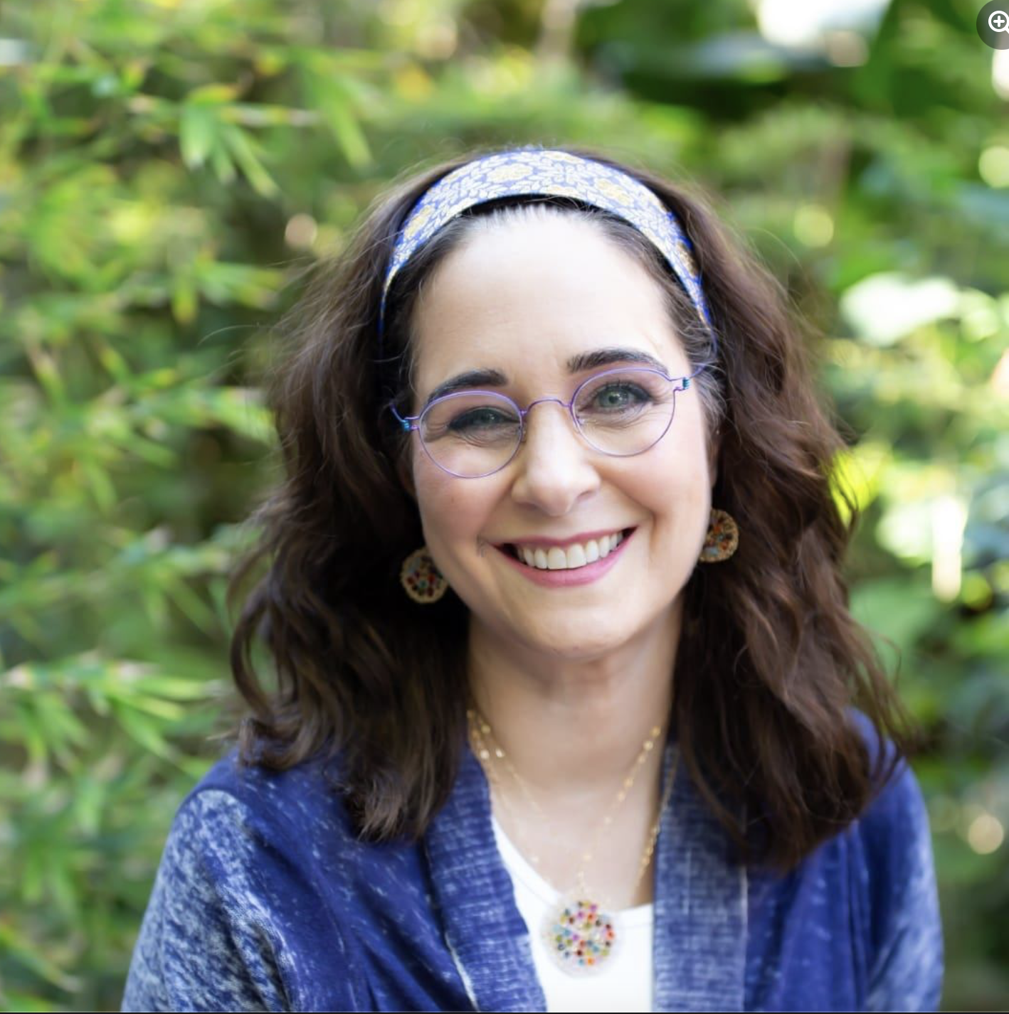 A smiling woman with curly brown hair, glasses, hoop earrings, and a blue patterned headband outdoors in a garden.