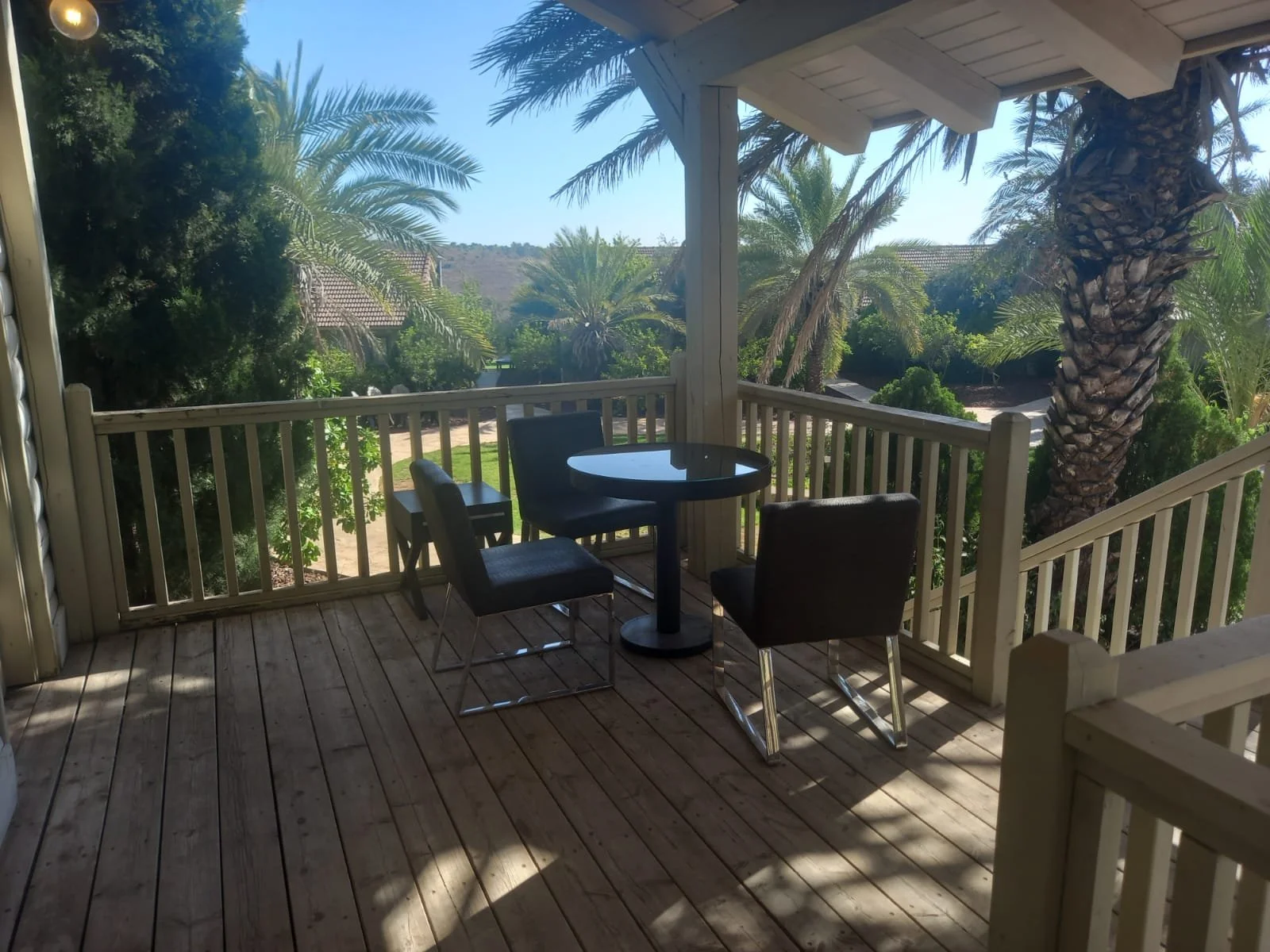 A wooden balcony with a small round table and four chairs, overlooking lush green trees and palm trees under a clear blue sky.