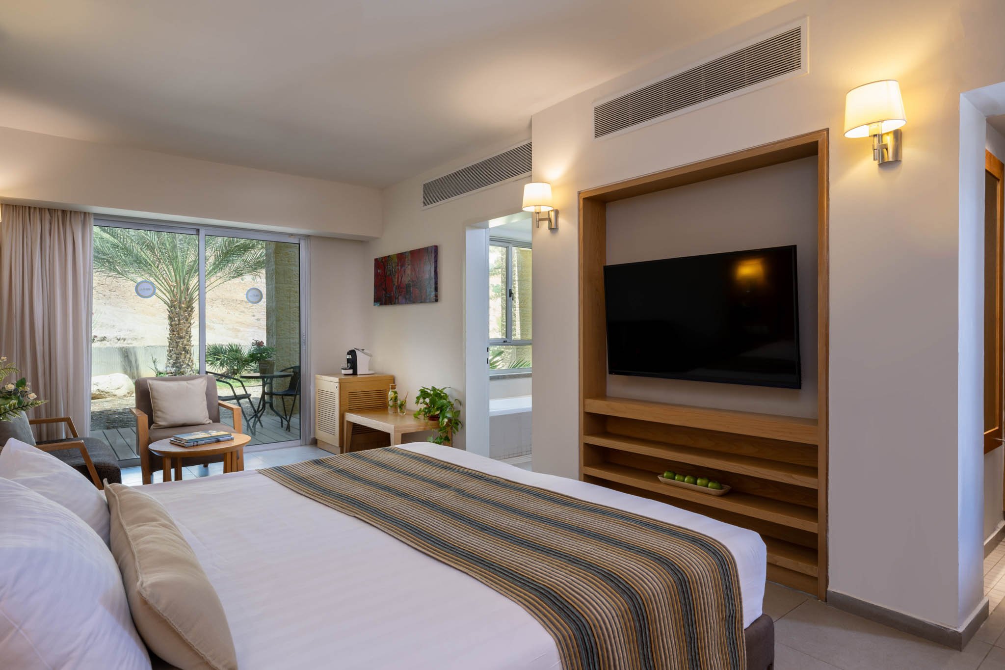 A hotel room with a large bed, two decorative pillows, and a beige headboard. There are two hanging pendant lights, a grey armchair, a small white round table with a flower arrangement, and a balcony with outdoor seating and a view of trees and buildings.