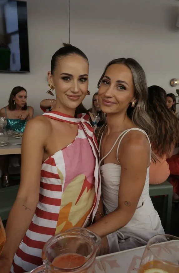 Two women sitting close together at a table, smiling at the camera, with several other women in the background at a social gathering or dinner event.