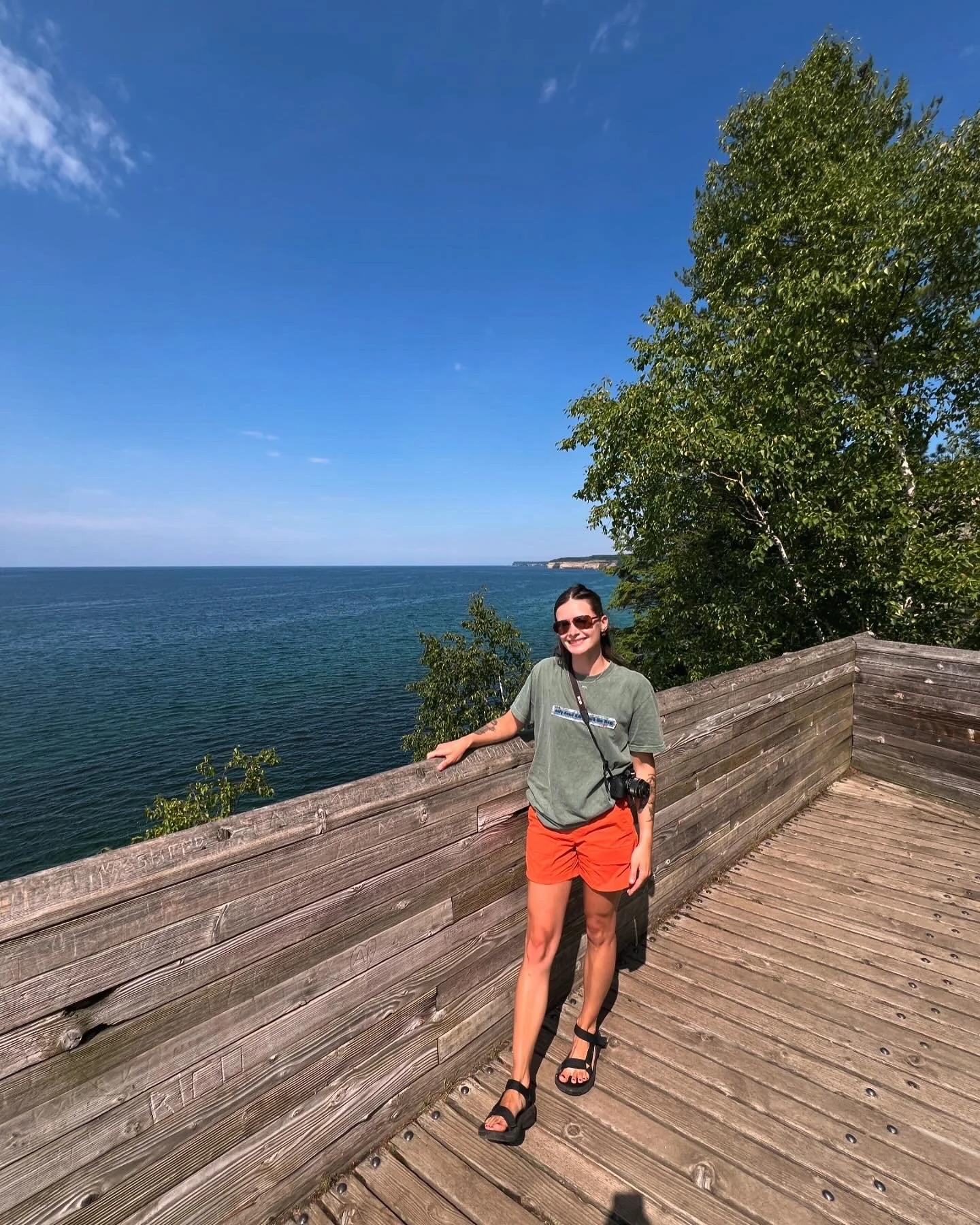 Woman with sunglasses, green t-shirt, and orange shorts standing on wooden observation deck by the ocean, with trees and cliffs in the background on a sunny day.