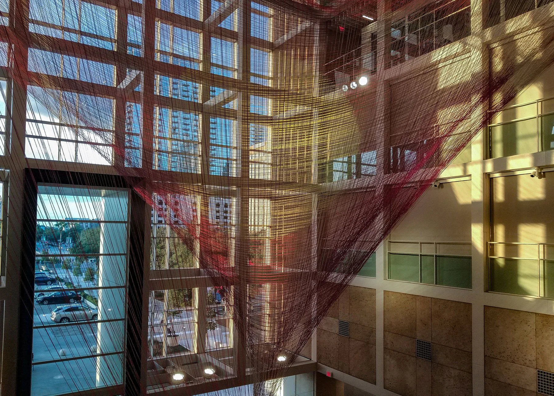 Interior of a modern building with large glass windows and a view of a parking lot outside. The ceiling features a colorful, netted art installation made of red and yellow threads.