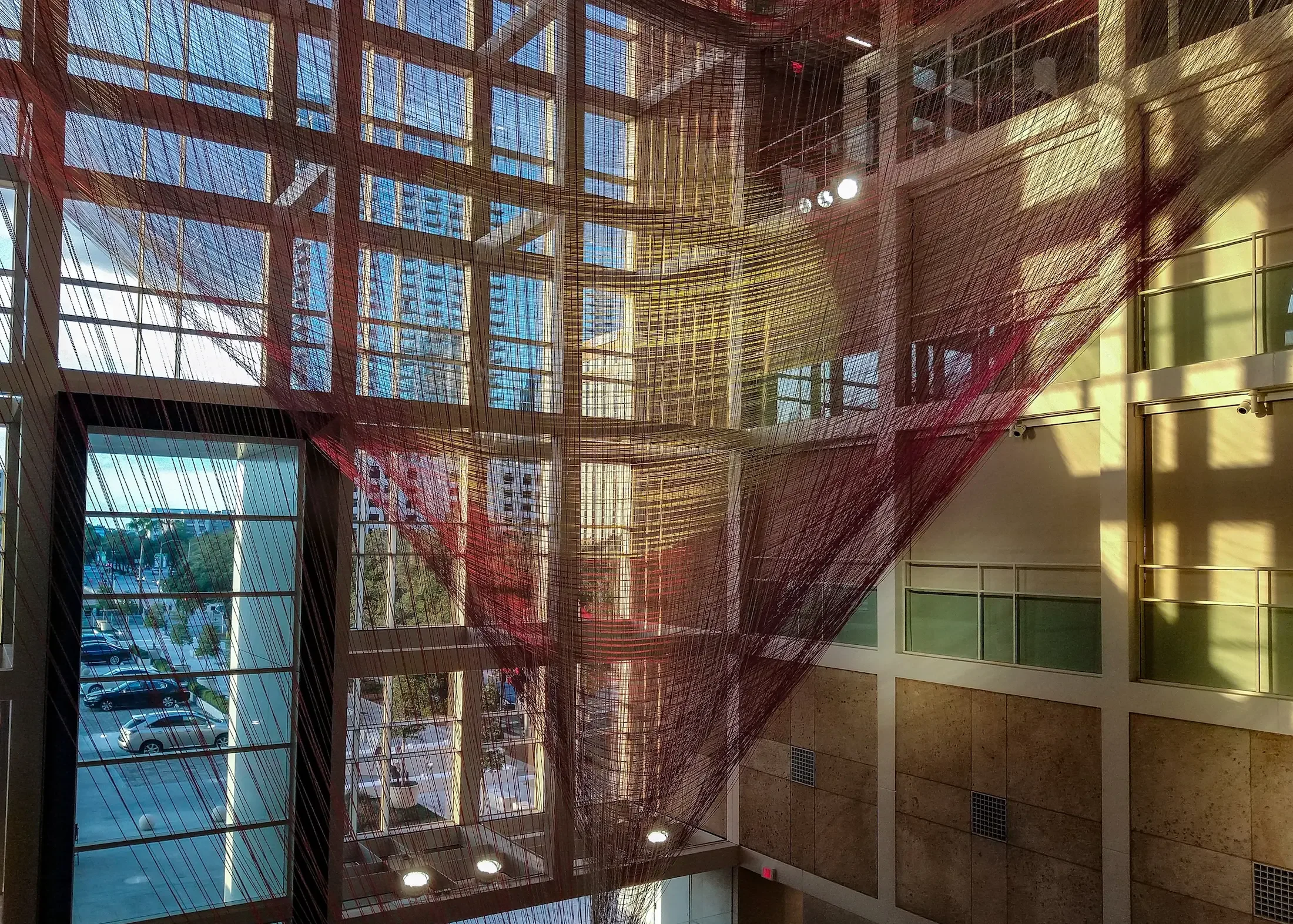 Interior of a modern building with large glass windows and a view of a parking lot outside. The ceiling features a colorful, netted art installation made of red and yellow threads.