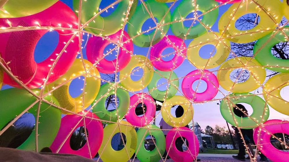 Colorful inflatable swim rings in yellow, pink, green, and blue, arranged on a metal frame with string lights, during dusk outdoors.