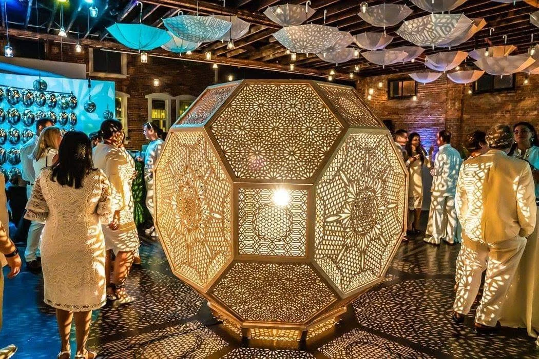 A large, geometric, lantern-like centerpiece with intricate cutout patterns, illuminated from within, casting patterned shadows. Surrounding people in formal attire are at a social gathering in a decorated brick venue with hanging umbrella decorations and blue wall accents.