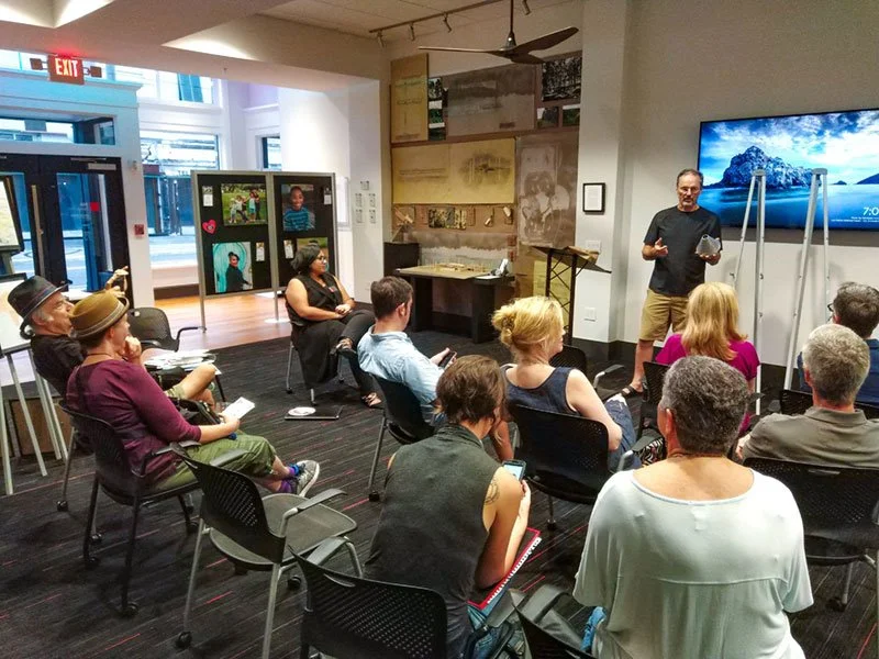 A group of people attending a presentation or lecture in a modern indoor space, with a man standing in front of a large screen, speaking to the audience.