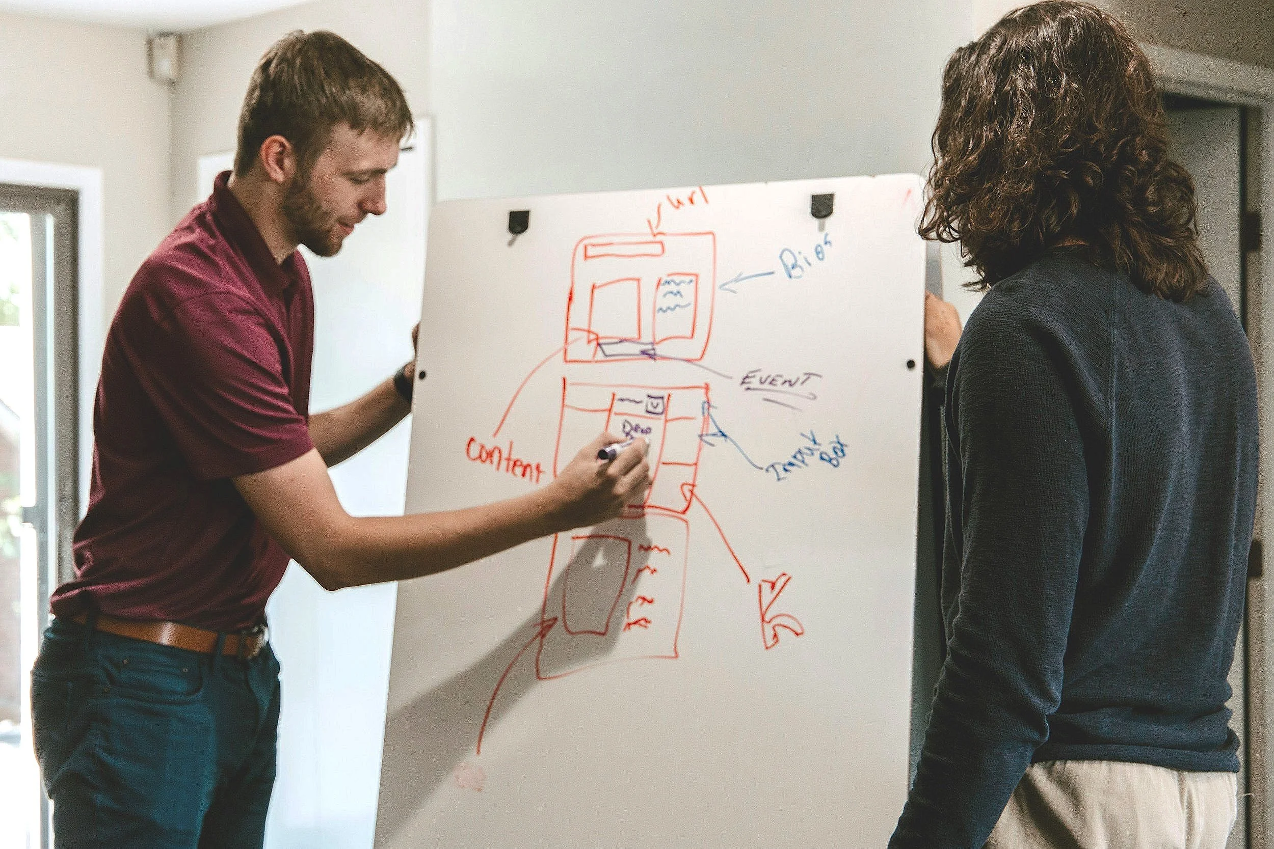 Two men are discussing a whiteboard with diagrams labeled 'Content,' 'Bio,' 'Event,' and 'Travel Box.' One man is writing on the board while the other observes.