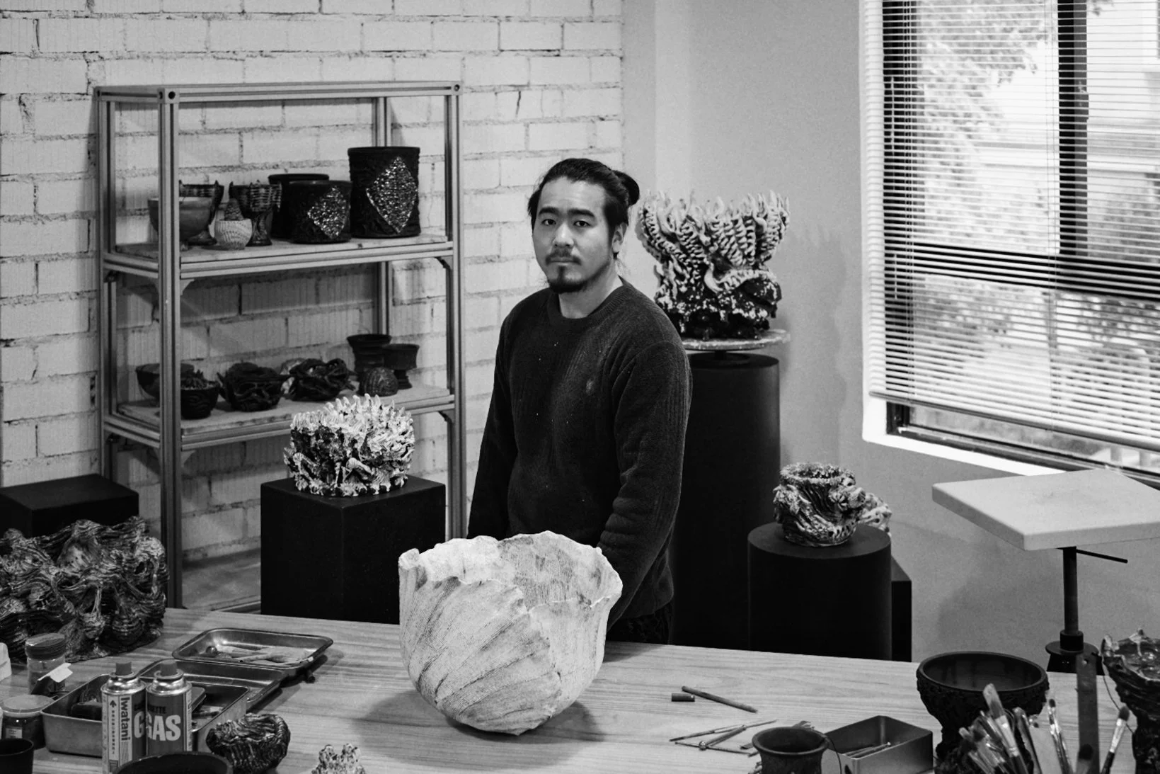 A man standing behind a table with various coral sculptures and tools, in an art studio with shelves and a window.