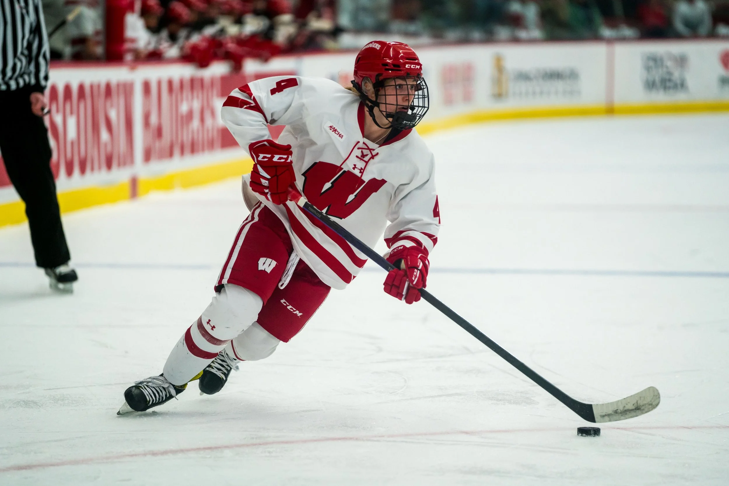 A hockey player in a white and red uniform with a Wisconsin Badgers logo on the chest, wearing a red helmet, gloves, and skates, is skating on the ice and handling a puck with a stick during a game.