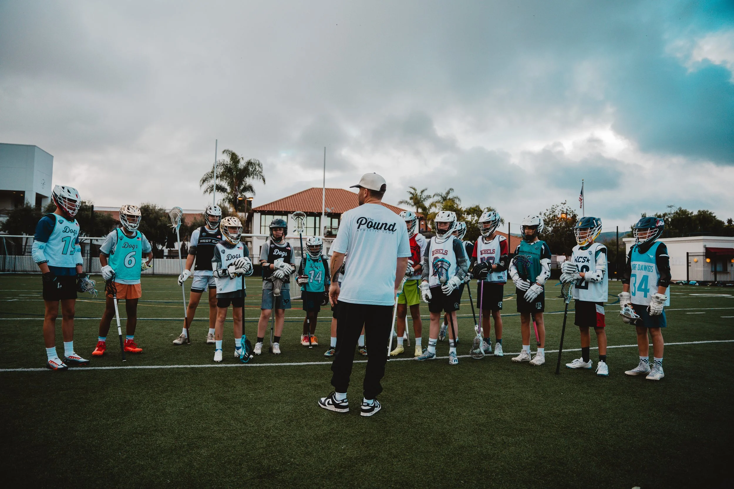 Lacrosse coach instructing a youth team on a field. The young players wear helmets, gloves, and jerseys with team names and numbers, holding lacrosse sticks, standing in a semi-circle around the coach under cloudy skies.